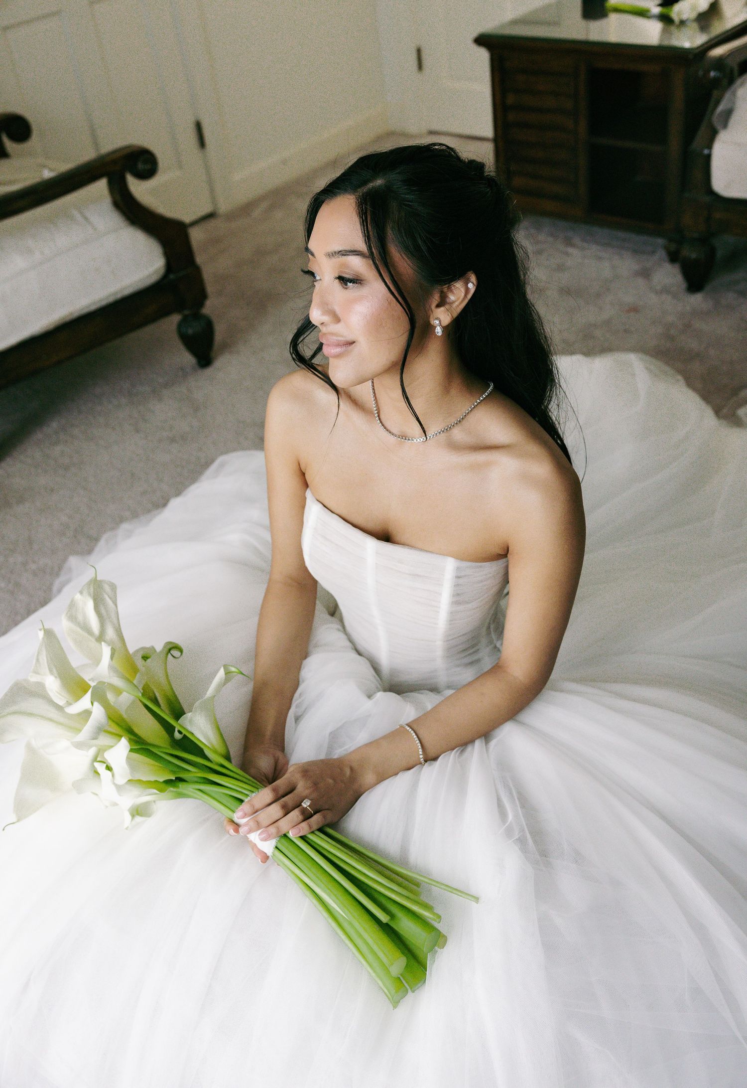 Bride in strapless white ballgown holding calla lily bouquet, sitting on floor in elegant room.