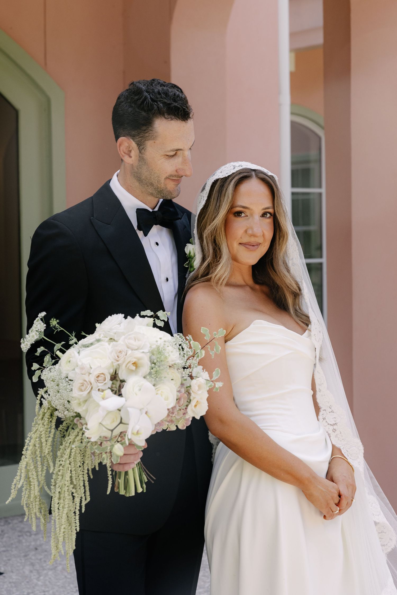 A bride in a white strapless gown and groom in black tuxedo pose together with an elegant white floral bouquet.