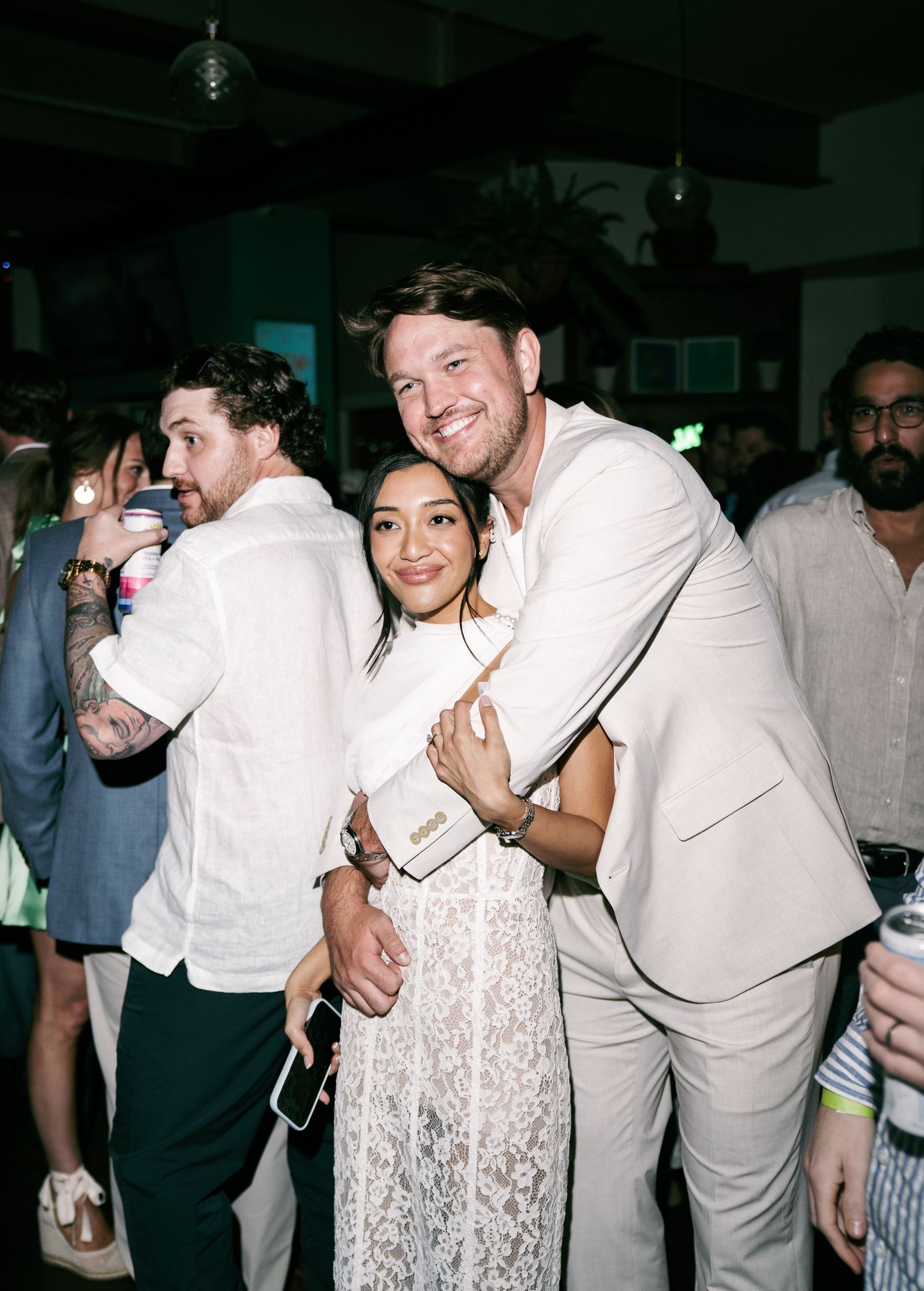 Couple hugging and smiling at a white party event, surrounded by guests in white outfits.