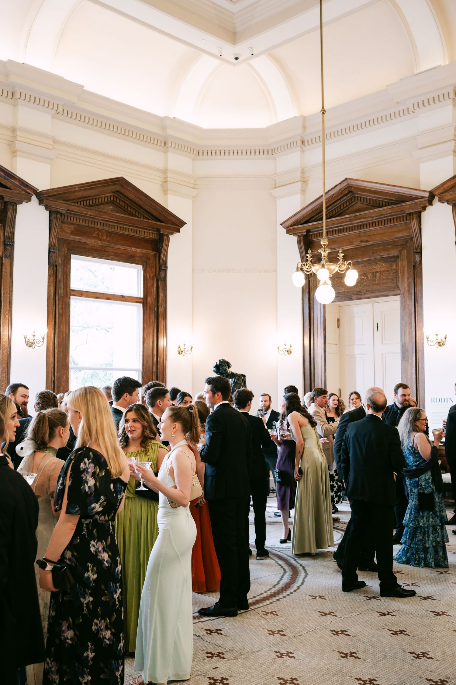 Wedding guests mingling in grand neoclassical venue with high ceilings and ornate architecture.