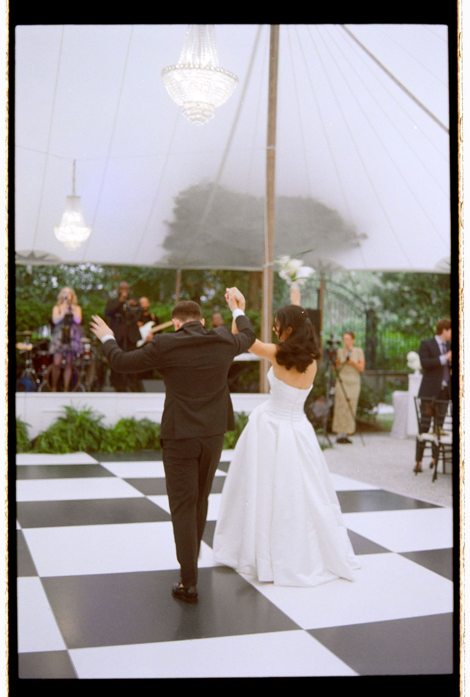 Bride and groom entering reception on black and white checkered dance floor under elegant tent.