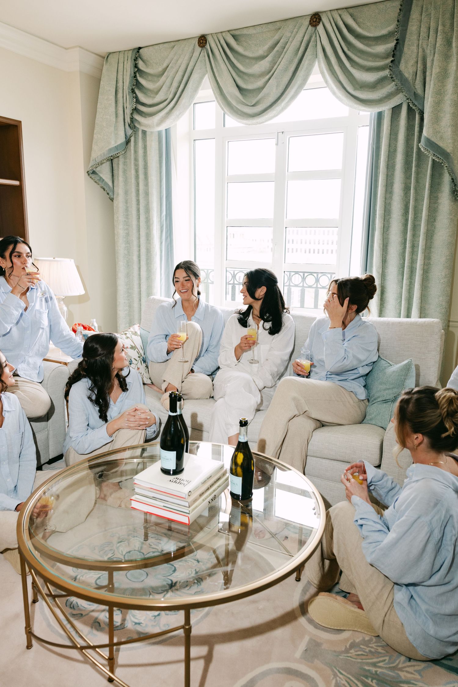 Group of bridesmaids in blue pajamas celebrating with champagne in an elegant hotel suite.