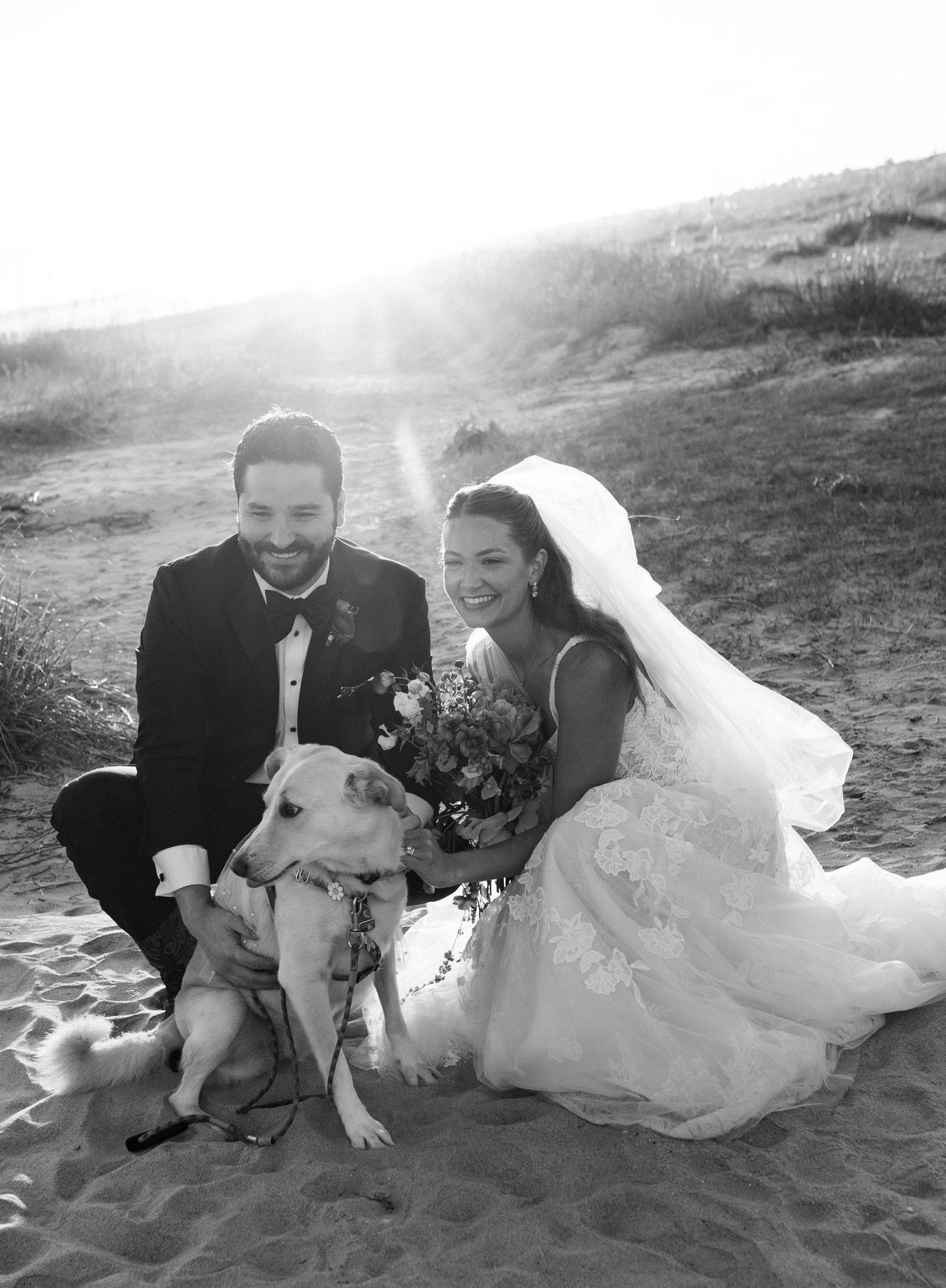 Smiling newlyweds pose with their dog on a sandy beach in black and white photo.