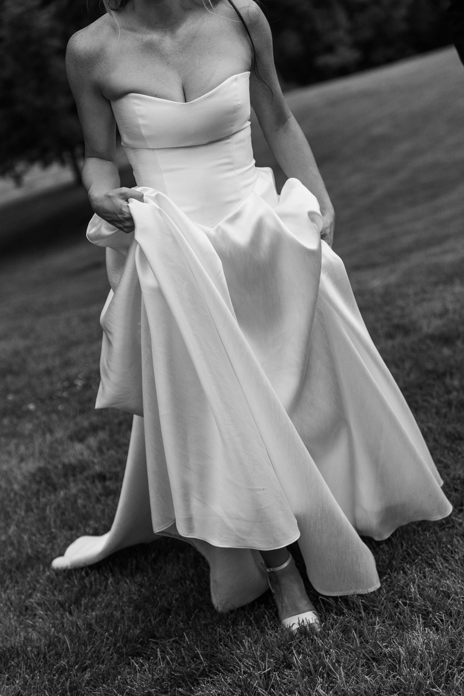 Black and white photo of bride in strapless satin gown lifting skirt on lawn.