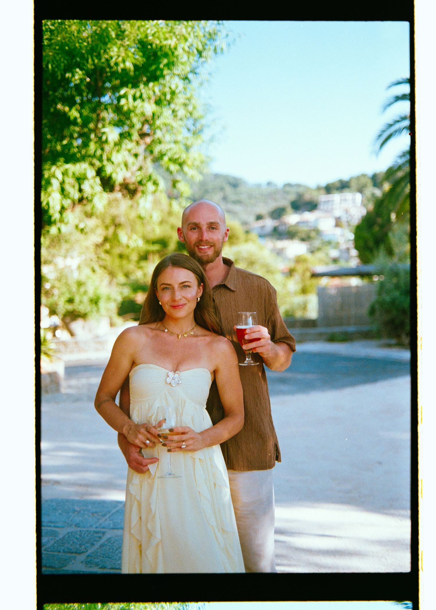 A couple smiles outdoors holding drinks, woman in white strapless dress, man in brown shirt, lush greenery background.