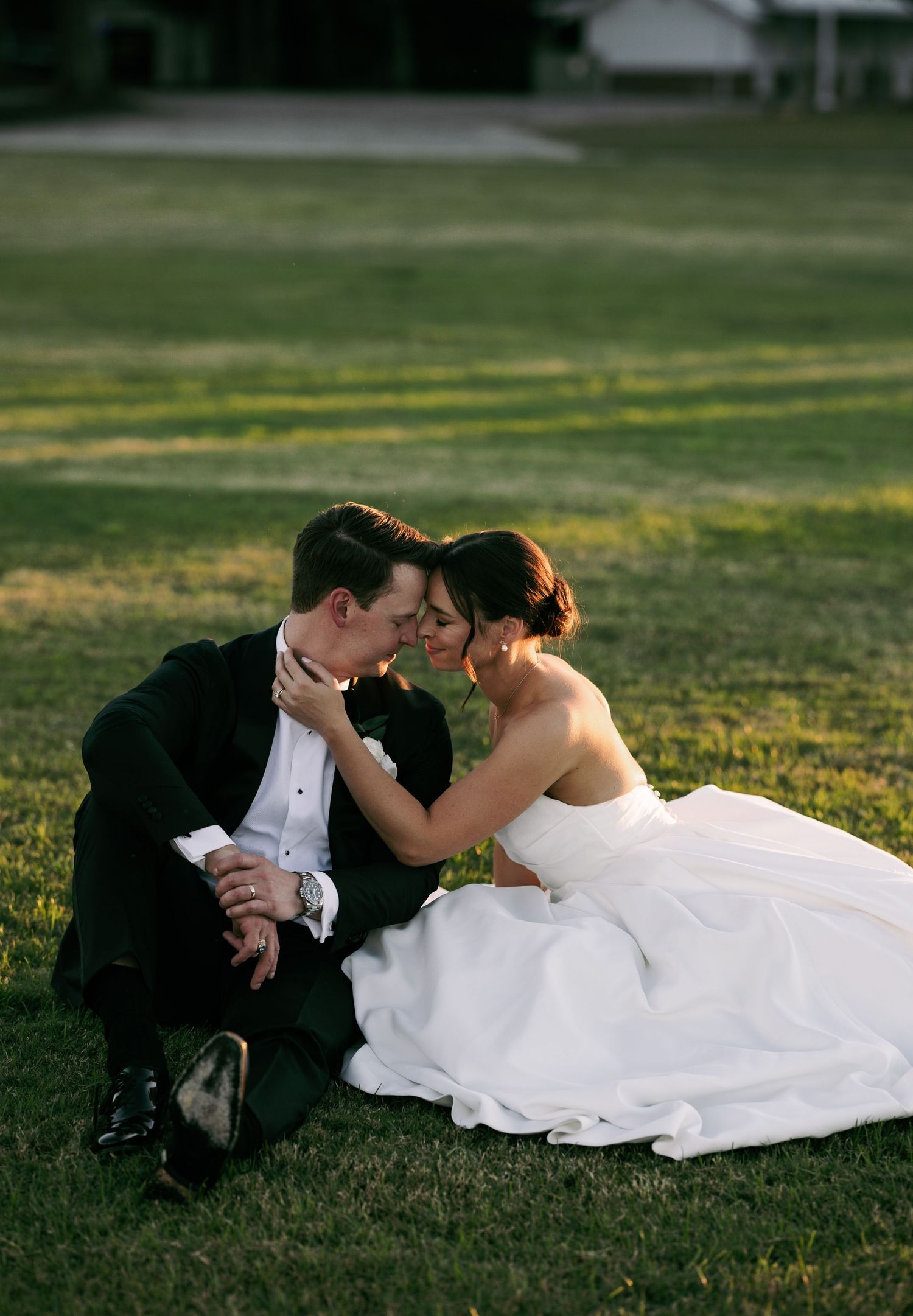 Bride and groom sitting on green grass, sharing a romantic moment in wedding attire, with the bride in a white gown.
