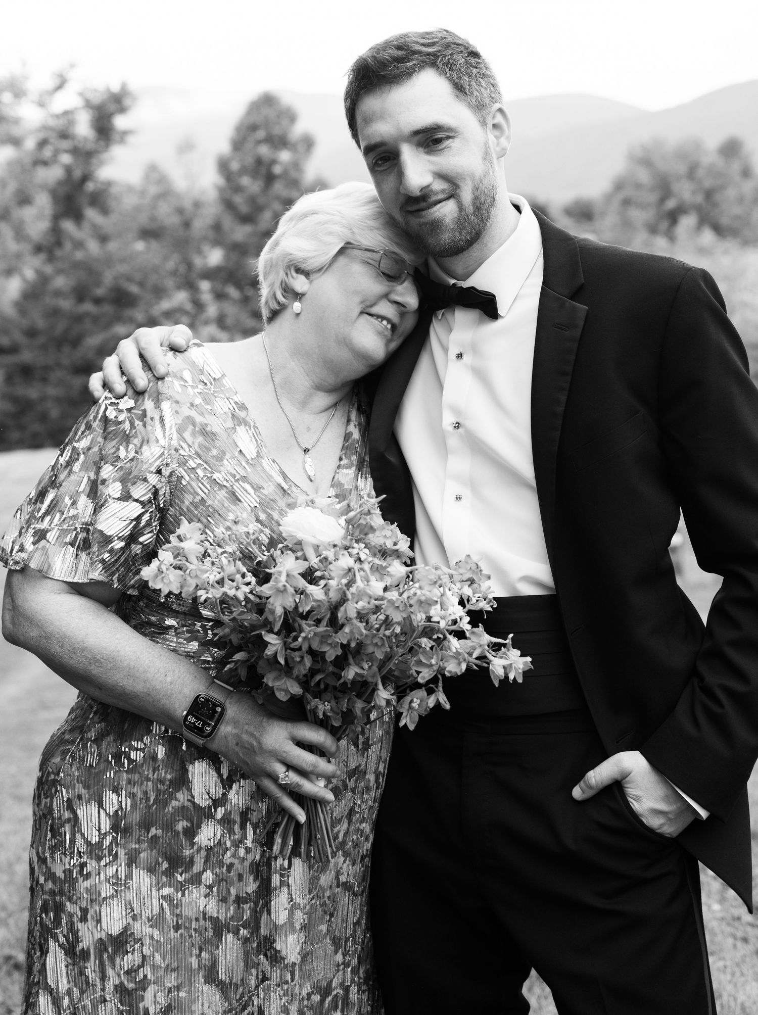 Groom in black tuxedo embracing older woman holding floral bouquet at outdoor wedding celebration.