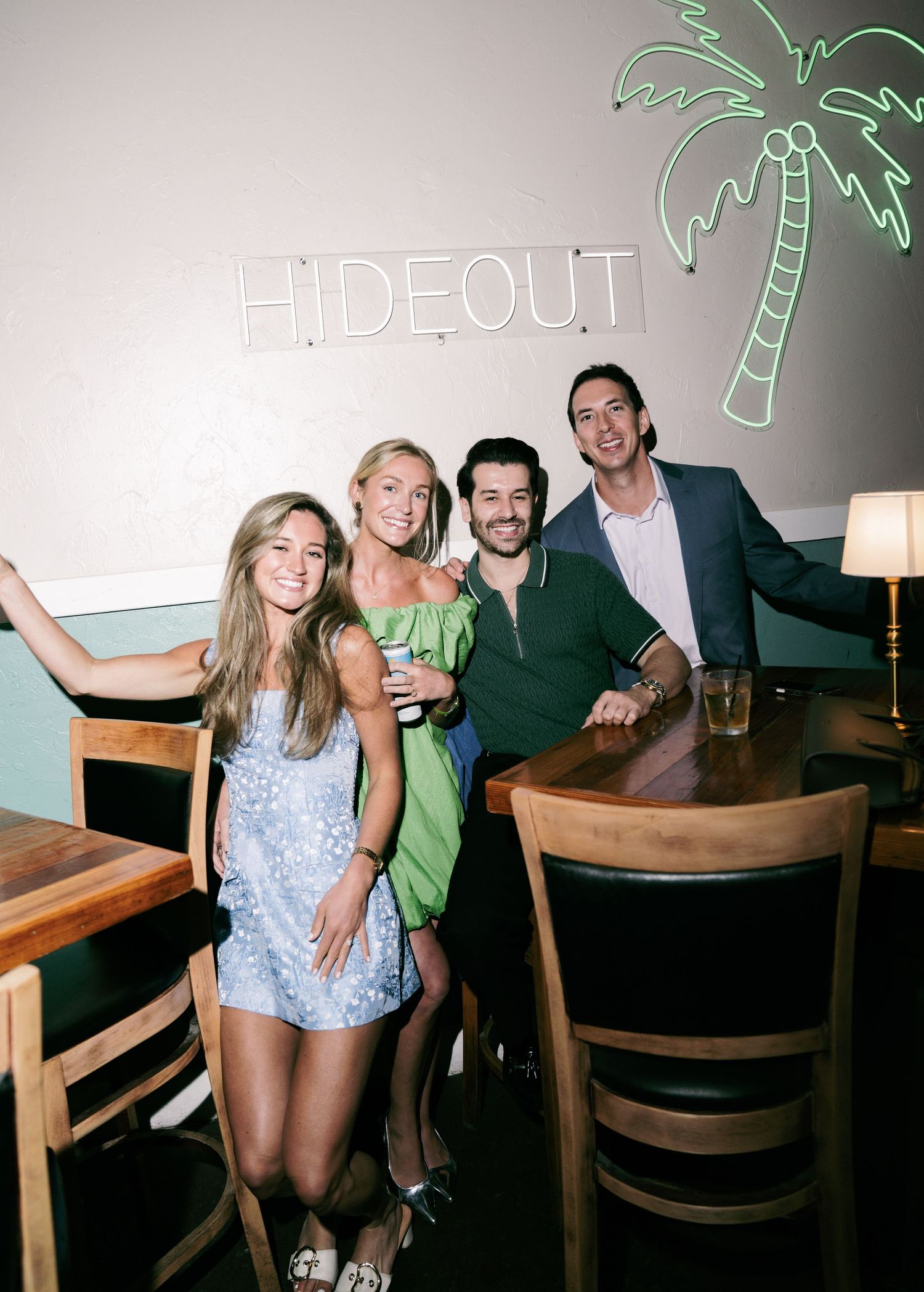Four friends smiling together at the Hideout bar, featuring neon signs and warm lighting.