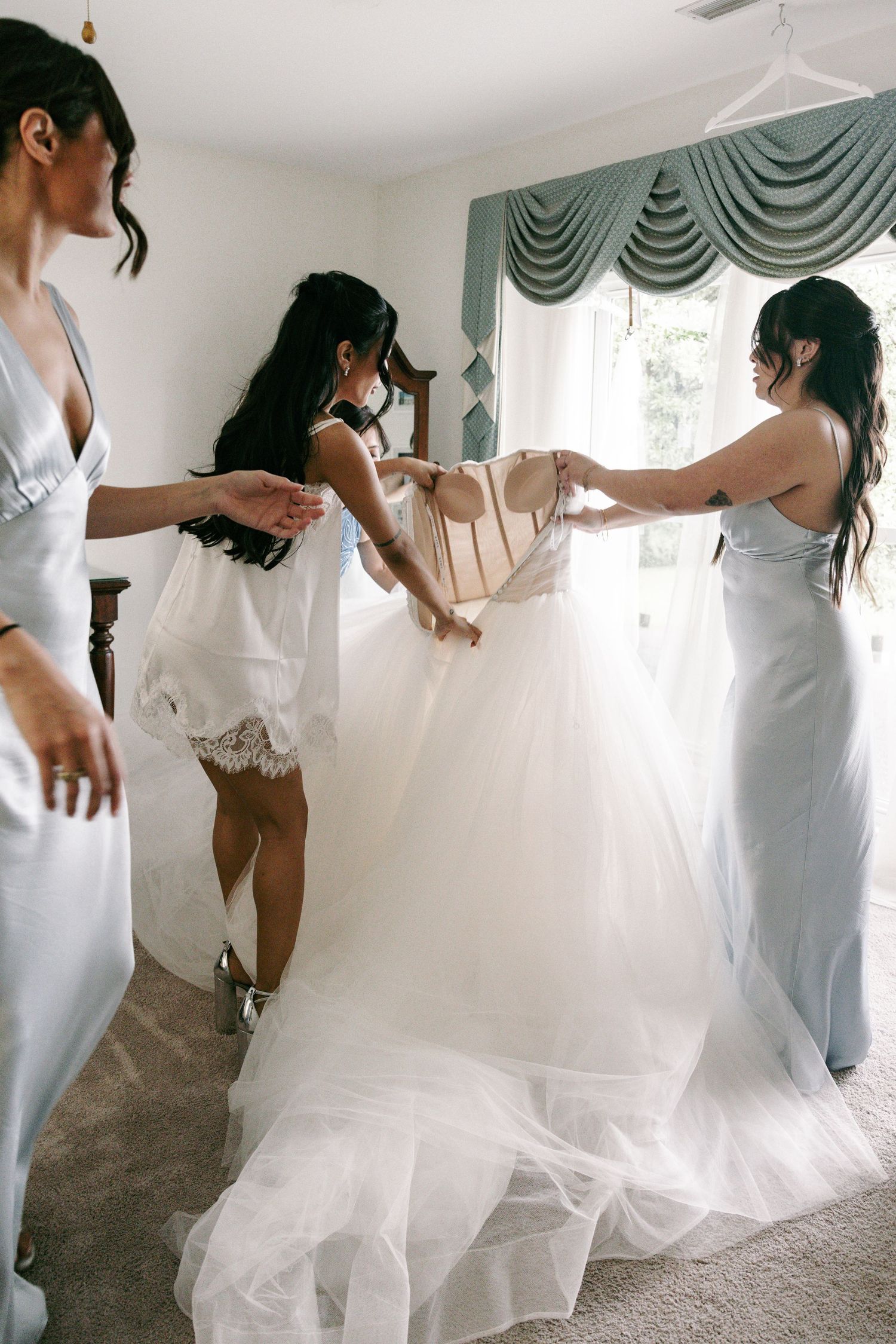 Bridesmaids in silver gowns help bride step into a flowing white tulle wedding dress in a bright bedroom.