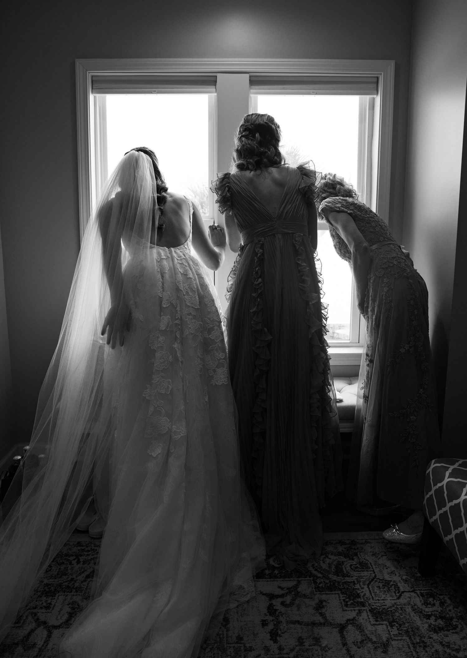 Black and white photo of bride in lace gown and veil with bridesmaids looking out window before wedding.