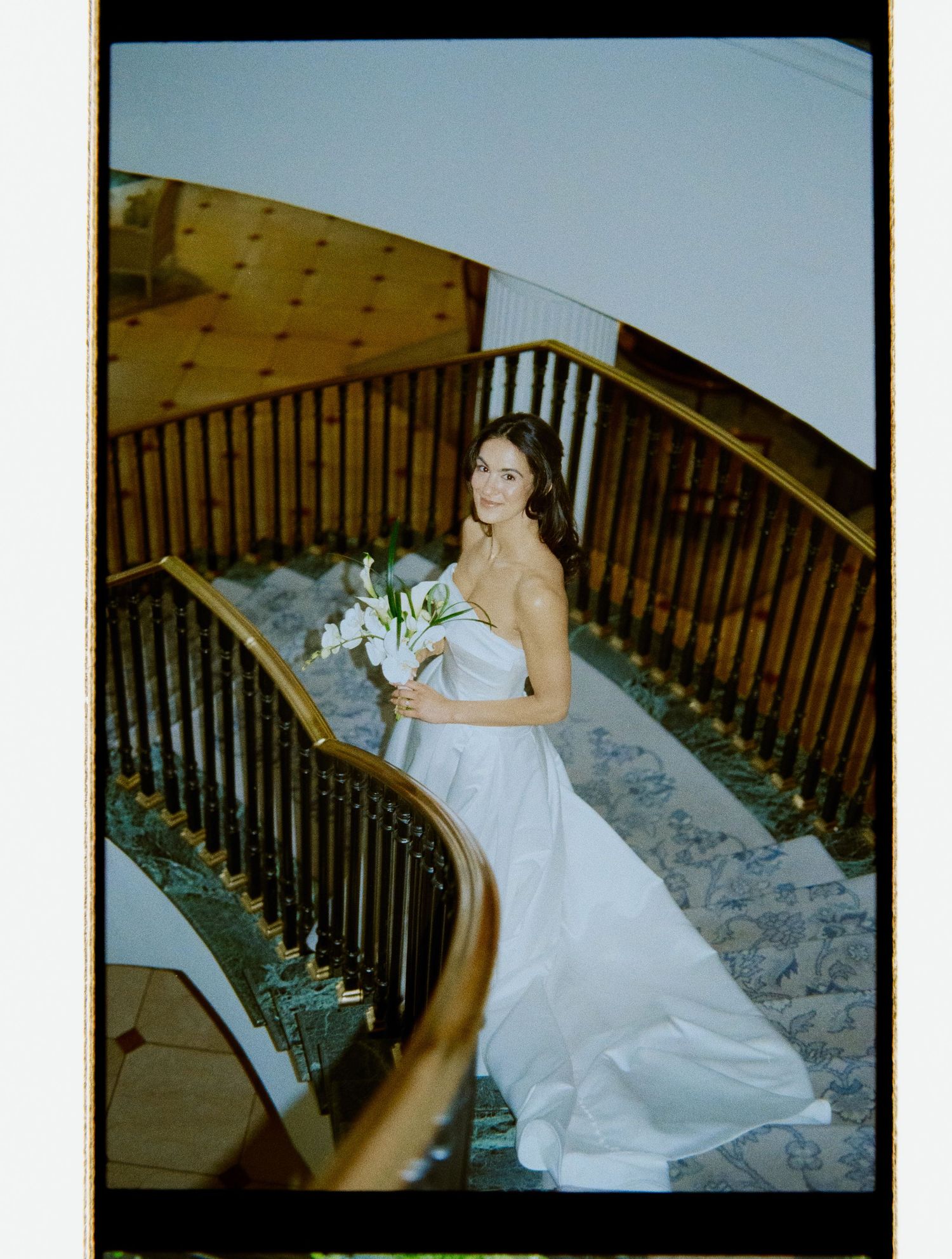 Bride in strapless white gown holding calla lily bouquet on grand staircase, film photo.