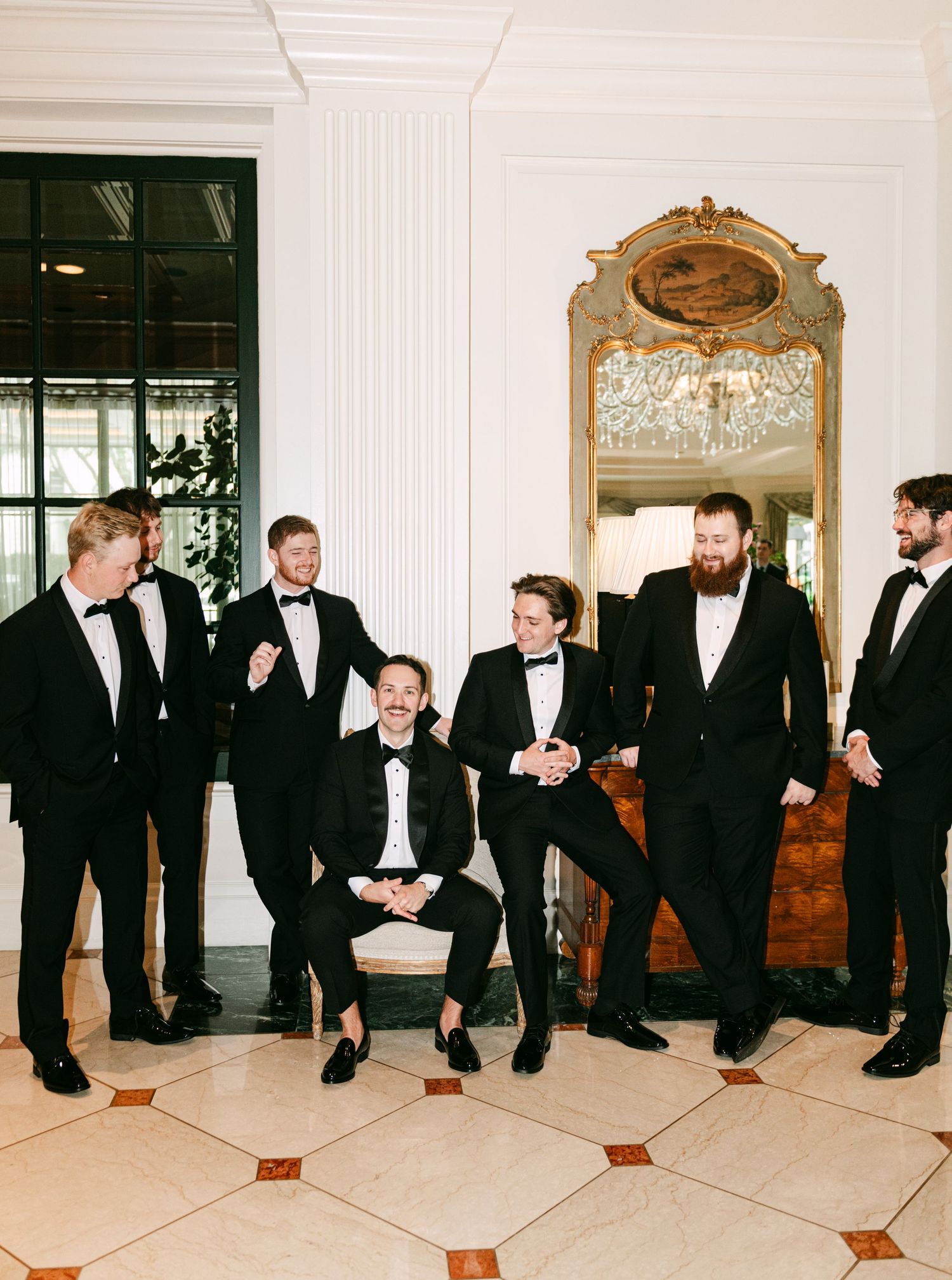 Seven groomsmen in black tuxedos laugh together in ornate wedding venue lobby.