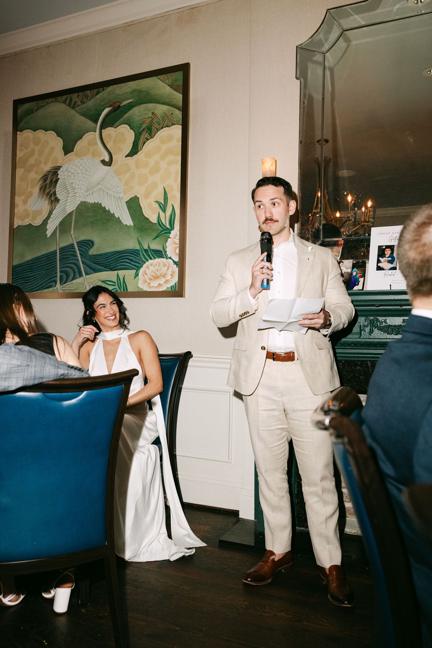 Man in cream suit giving a wedding speech with microphone, bride seated nearby, elegant venue with crane artwork.