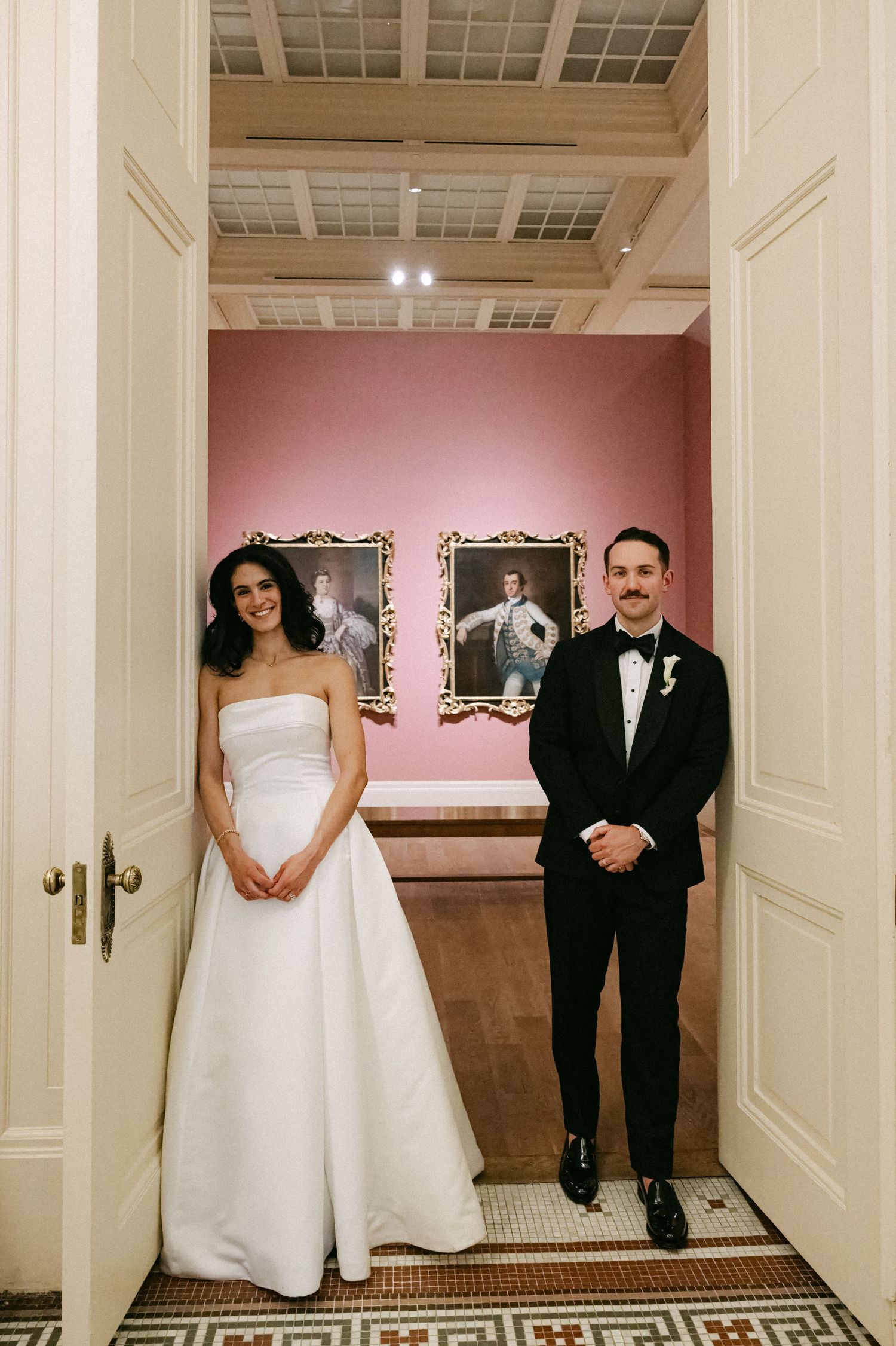Bride in strapless white gown and groom in tuxedo posing in art museum doorway.