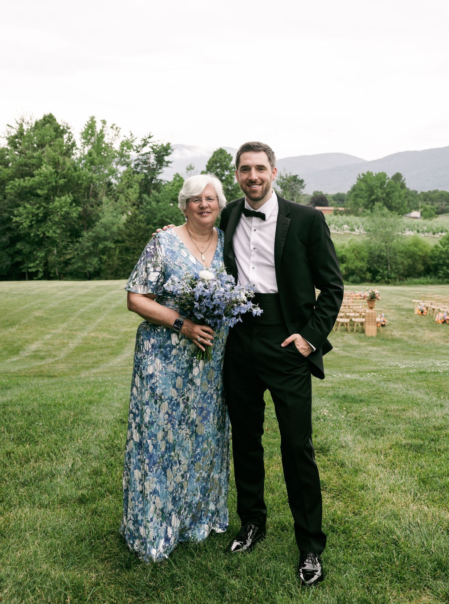 Groom in black tuxedo poses with older woman in blue floral dress holding bouquet at outdoor wedding.