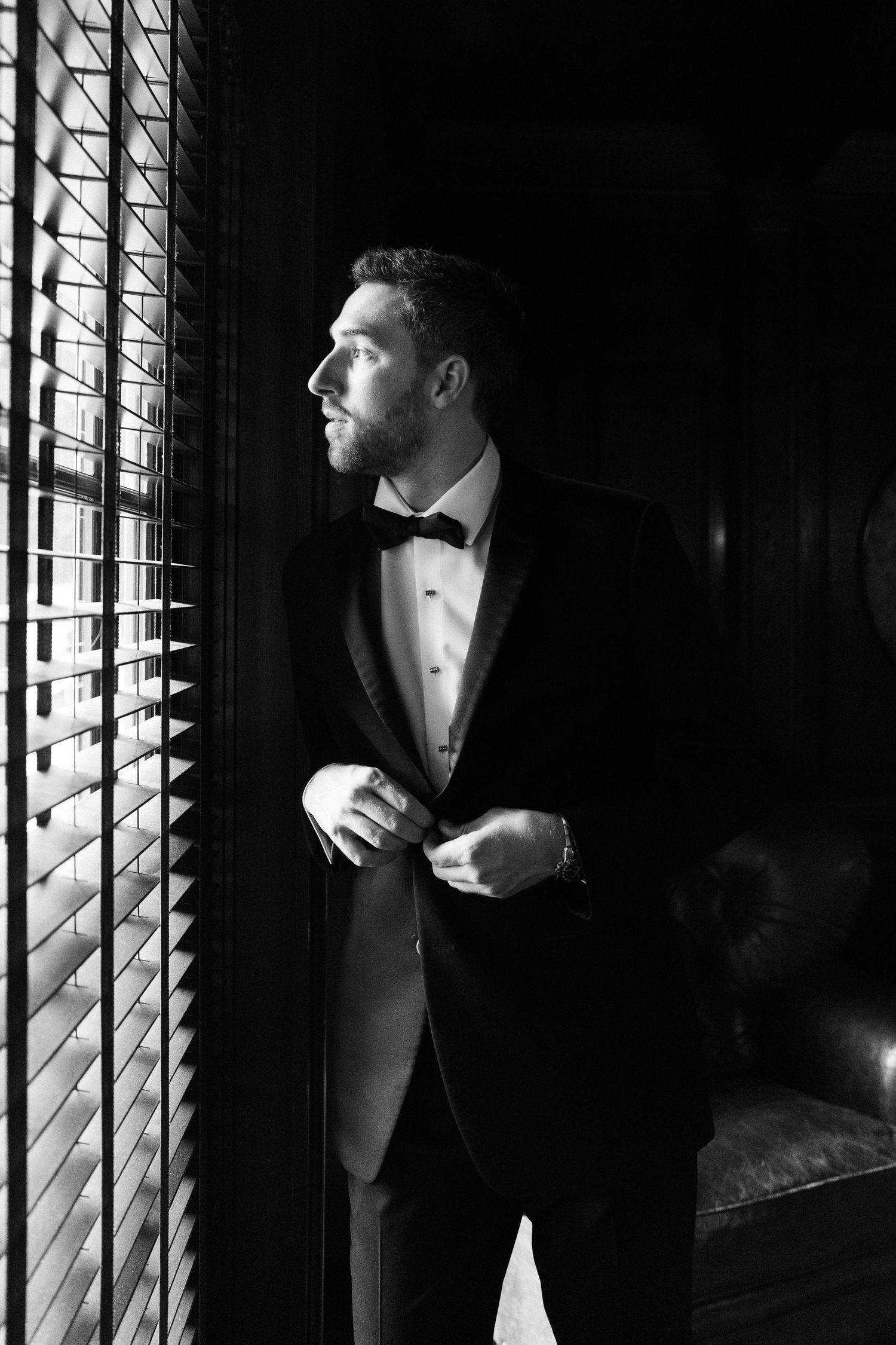 Dapper groom in black tuxedo and bow tie gazes out window in dramatic black and white portrait.