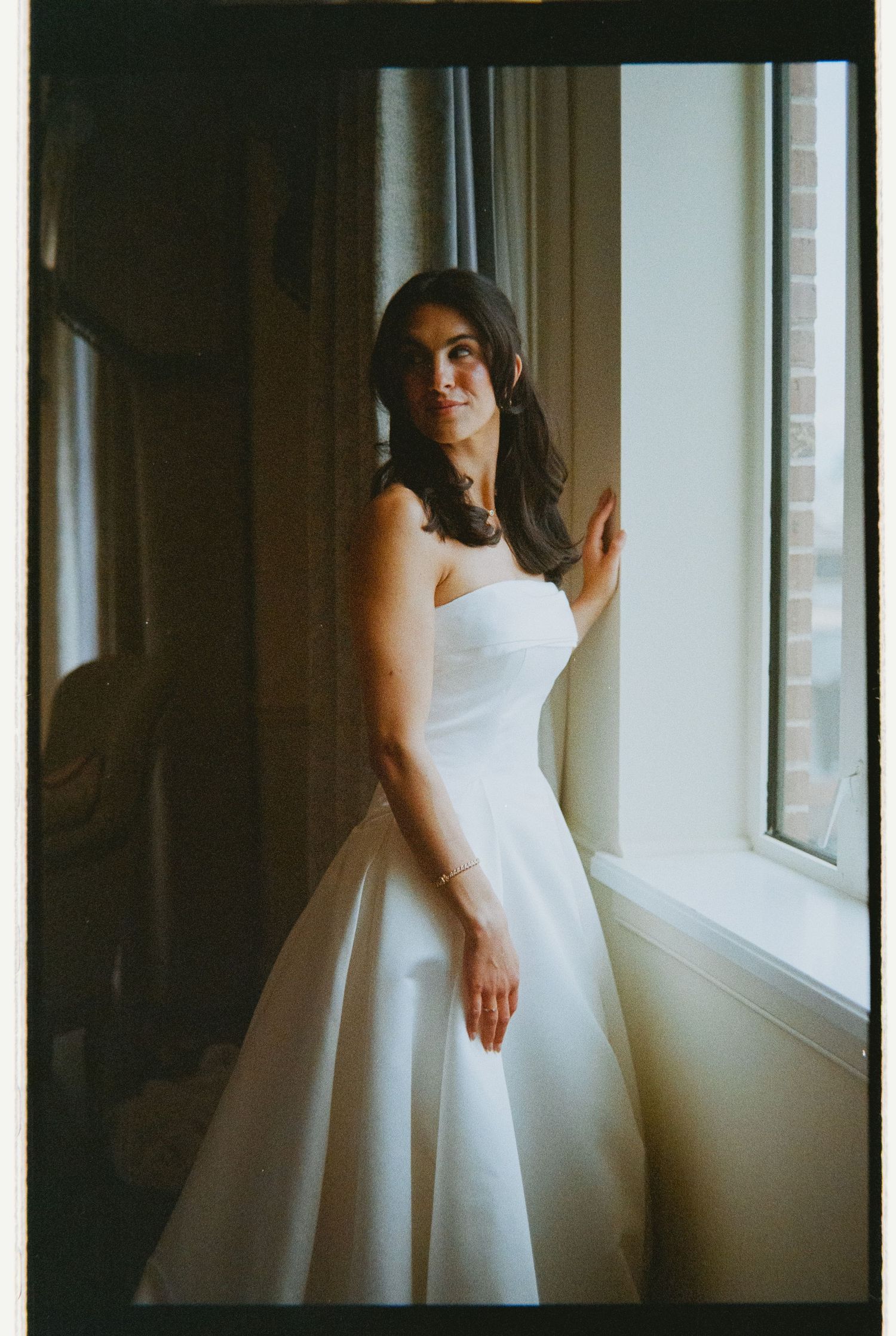Bride in strapless white ball gown poses elegantly by tall window in soft natural light, film photo.