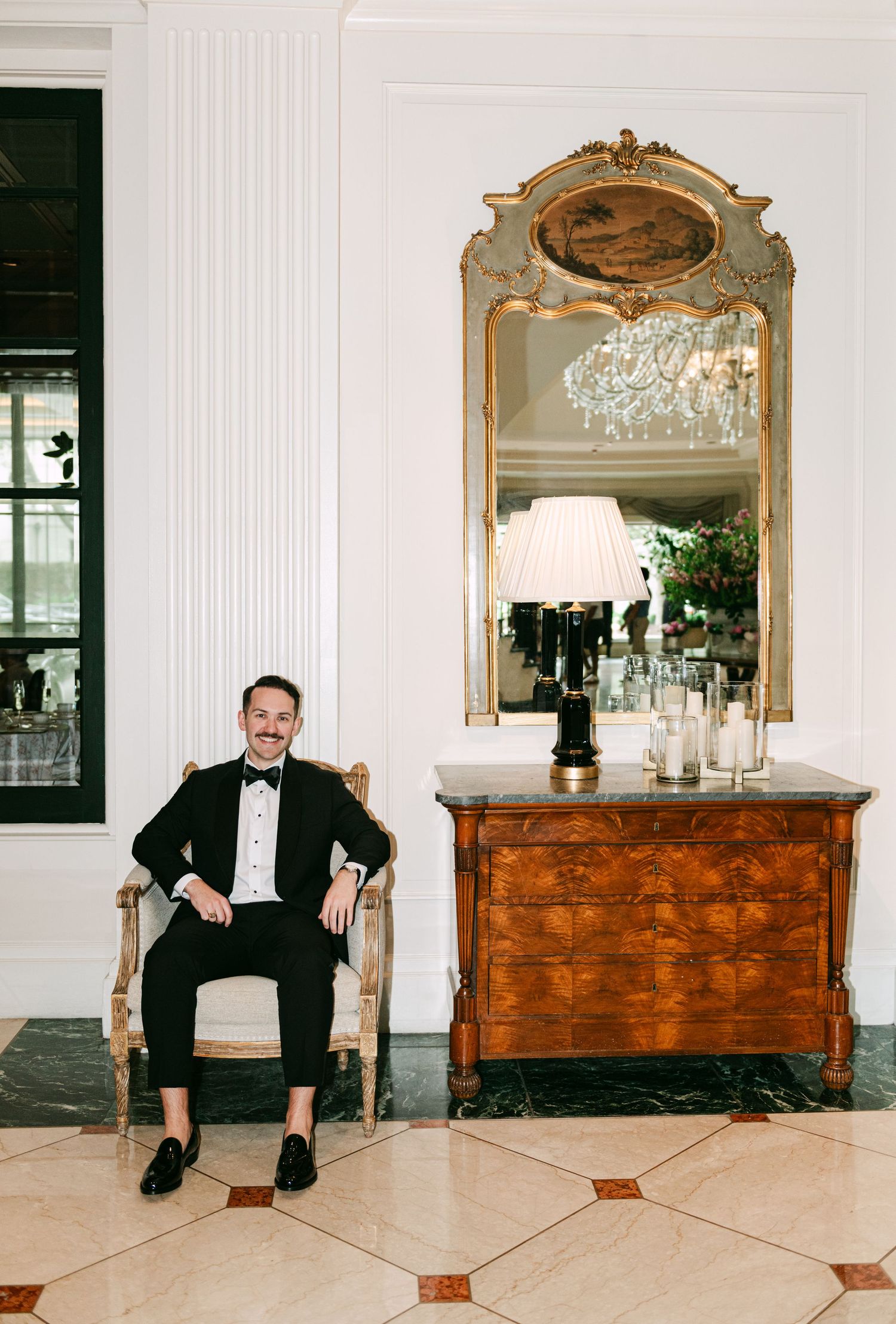 Dapper groom in black tuxedo sitting in ornate chair beside antique dresser with gilded mirror.