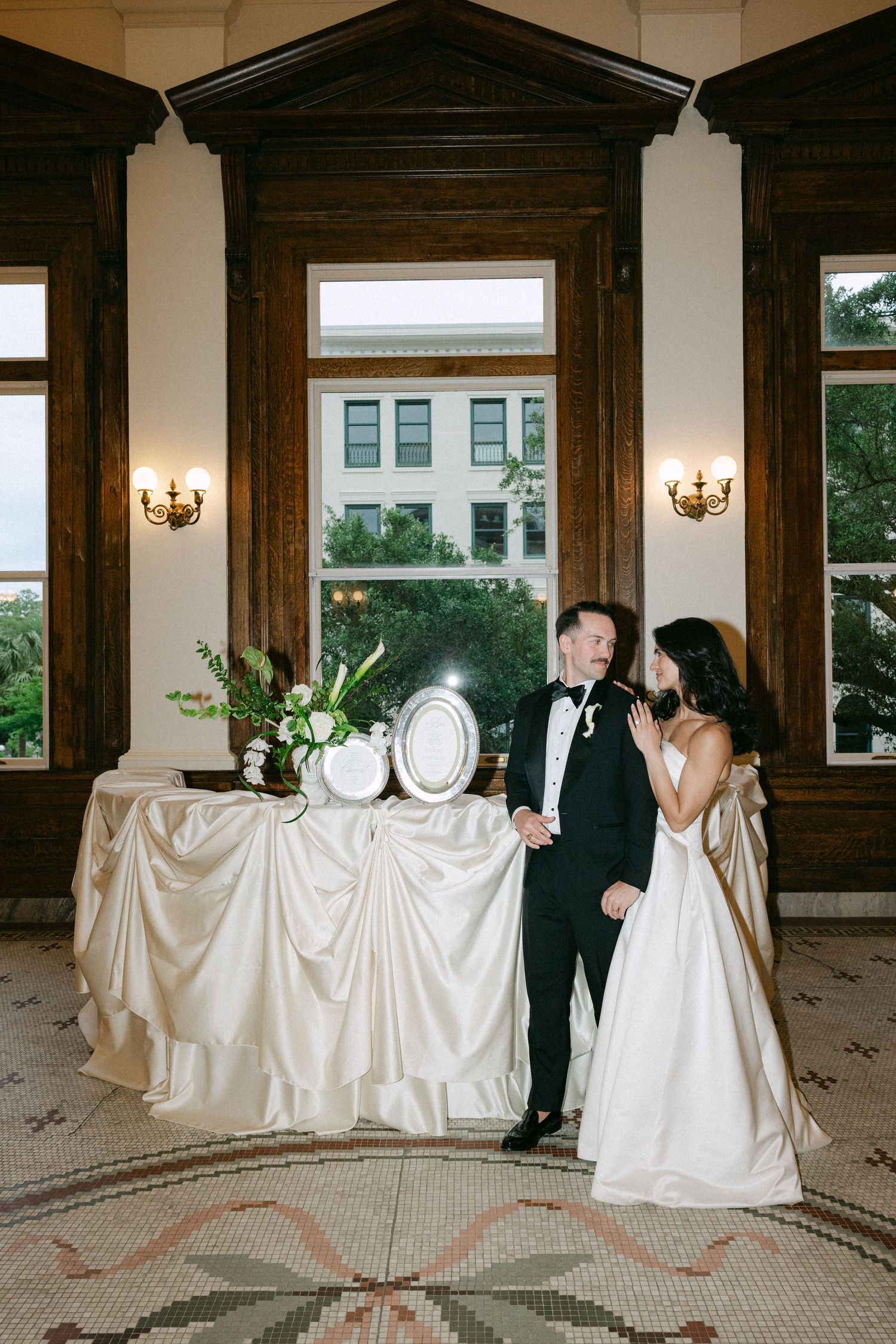 Bride and groom standing beside elegant ivory satin wedding cake table in ornate wood-paneled venue interior.