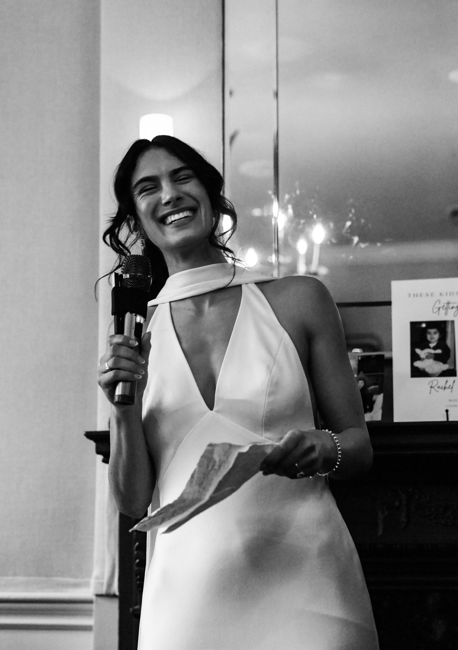 Bride in white halter dress laughing while giving speech at wedding reception.