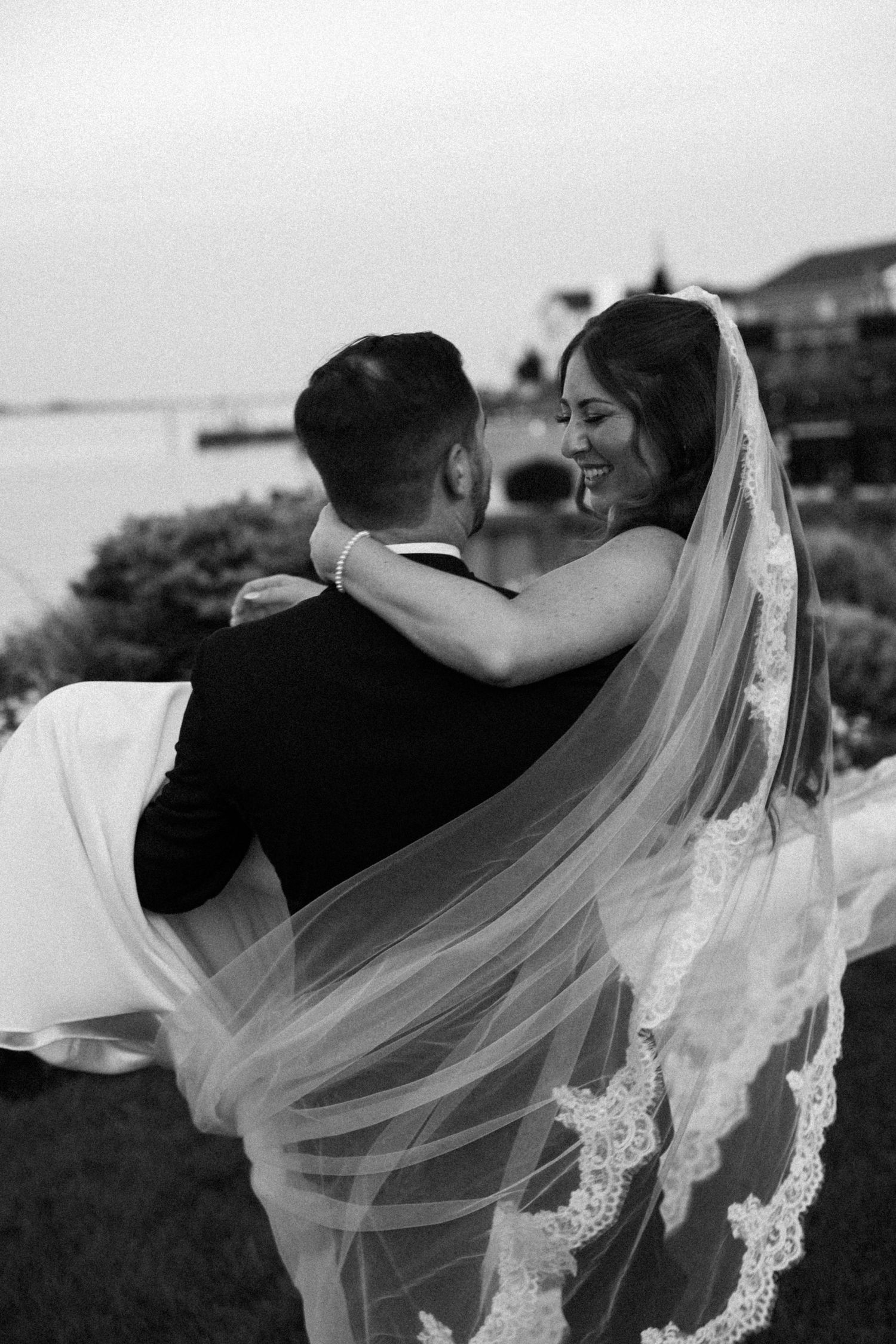 Groom carrying bride in lace veil outdoors near waterfront in black and white wedding photo.