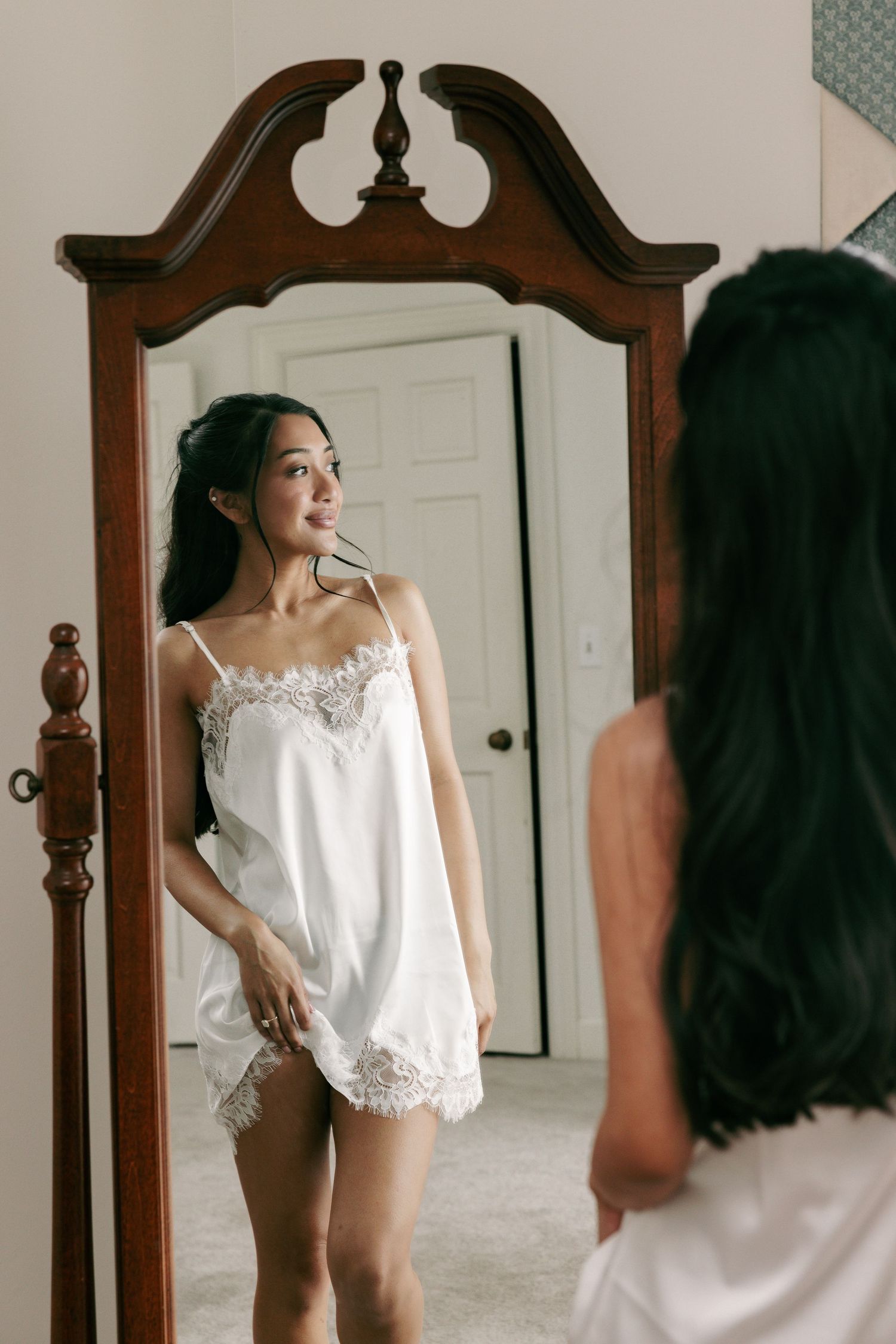 Bride in white lace satin slip dress admiring herself in an ornate wooden floor mirror before her wedding.