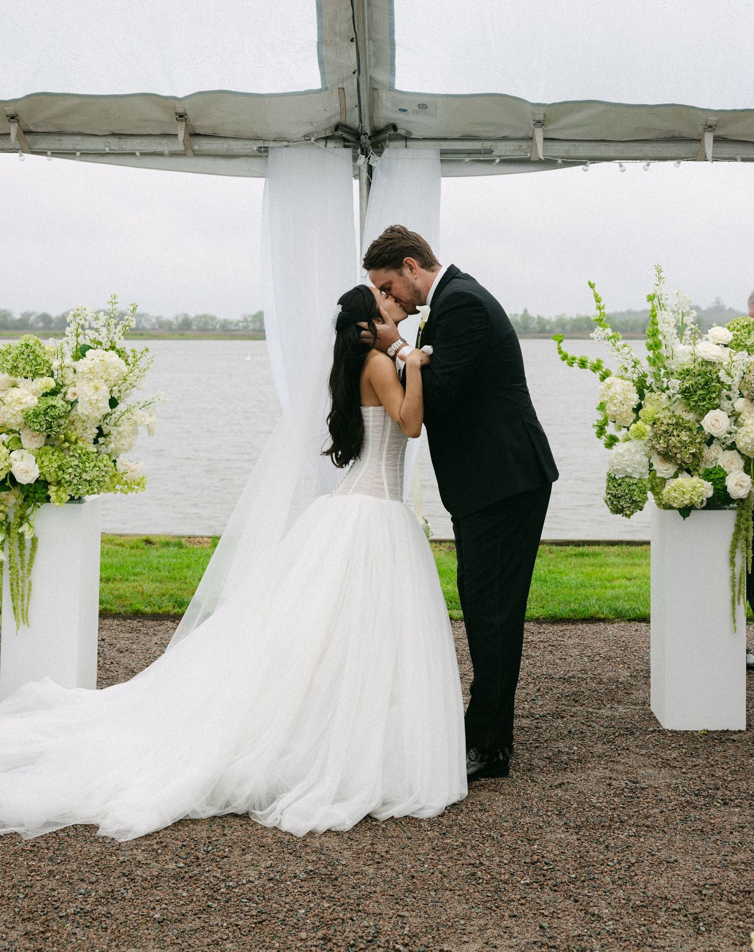 Bride and groom sharing a first kiss under a tent altar with white floral arrangements by a lake.