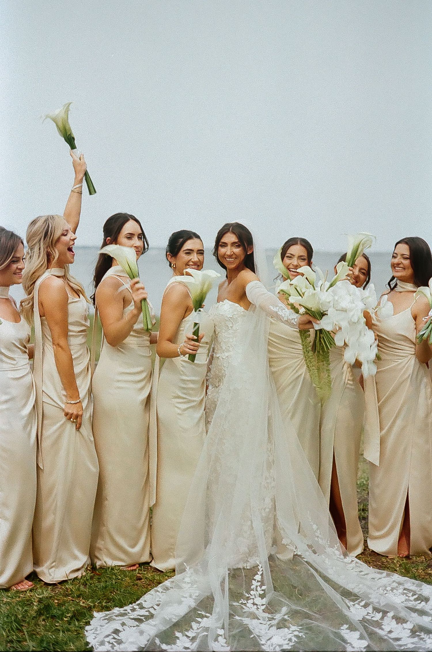 Bridesmaids in champagne dresses pose playfully in a line holding white bouquets.