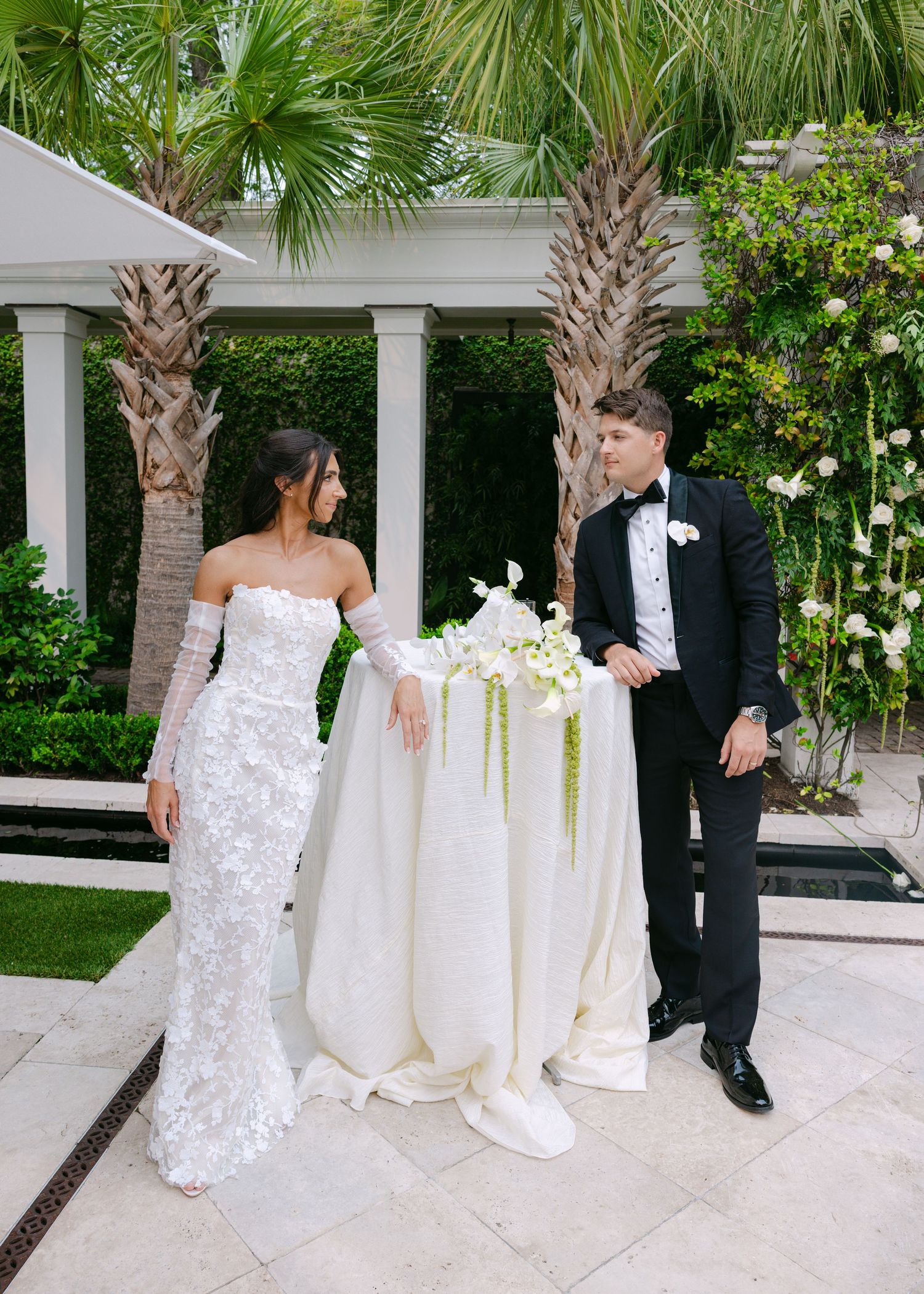 A wedding couple and guests stand beneath white columns and palm trees at an elegant outdoor venue.