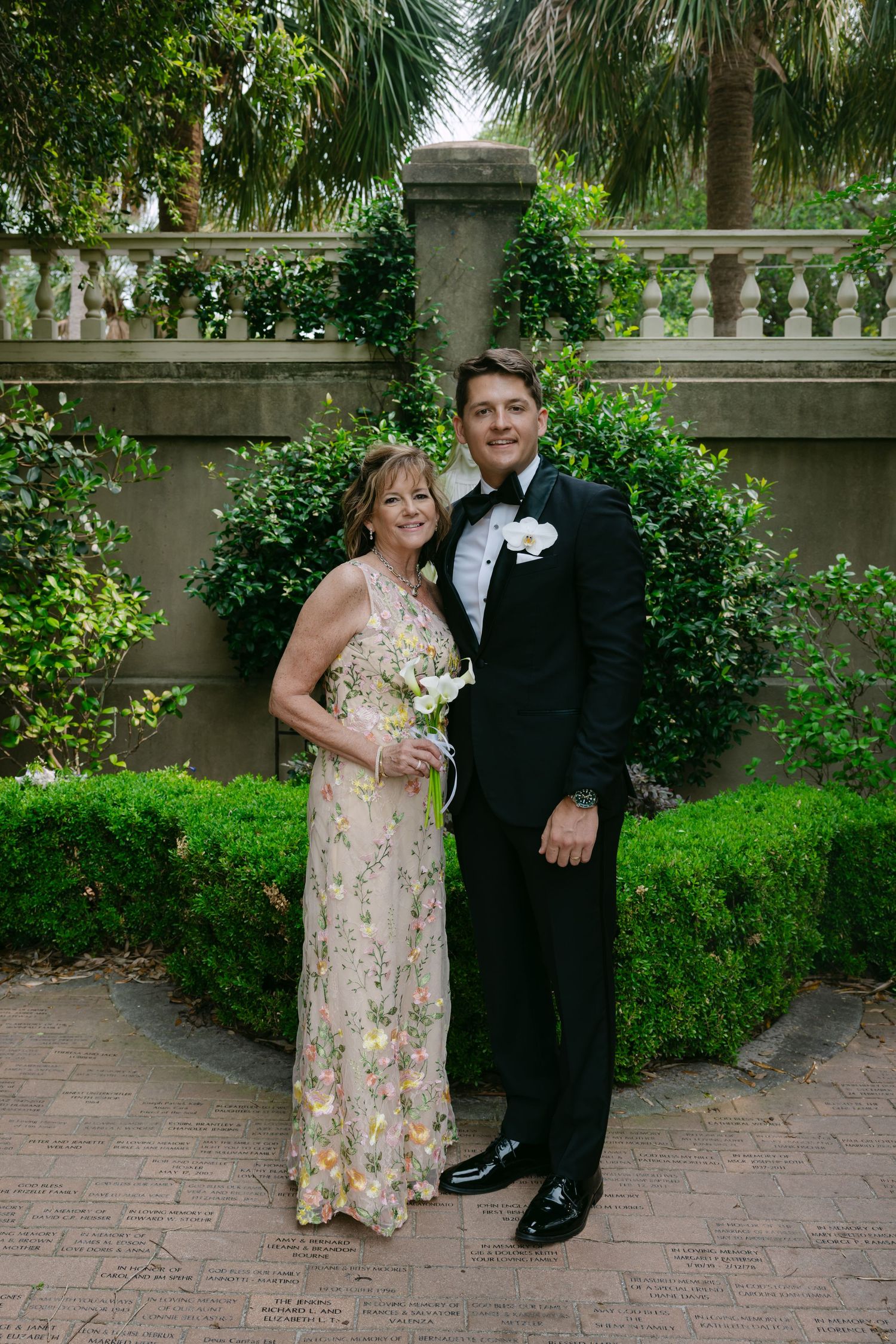 Wedding guests in colorful attire pose on brick pathway with garden backdrop.