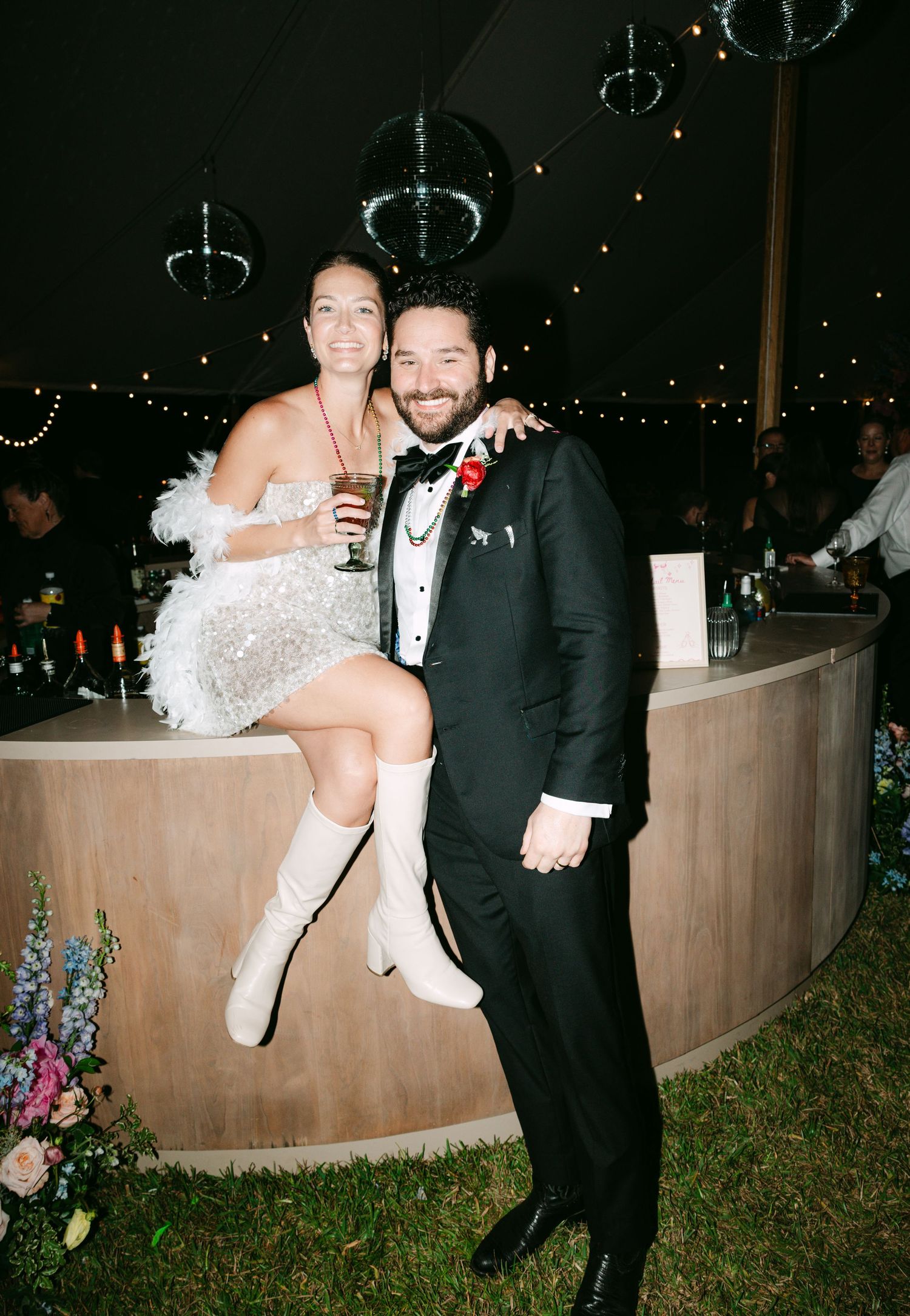 Bride in feathered mini dress and groom in tuxedo posing at wedding reception bar.