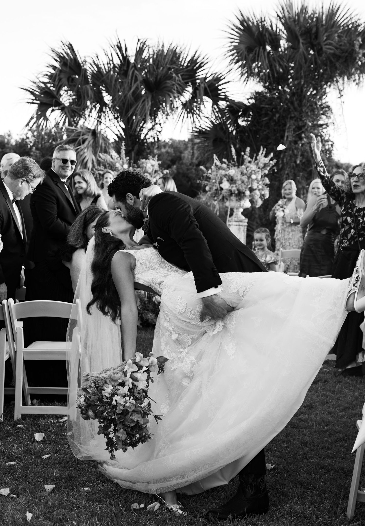 Black and white photo of bride and groom sharing a romantic dip kiss at outdoor wedding ceremony with guests watching.