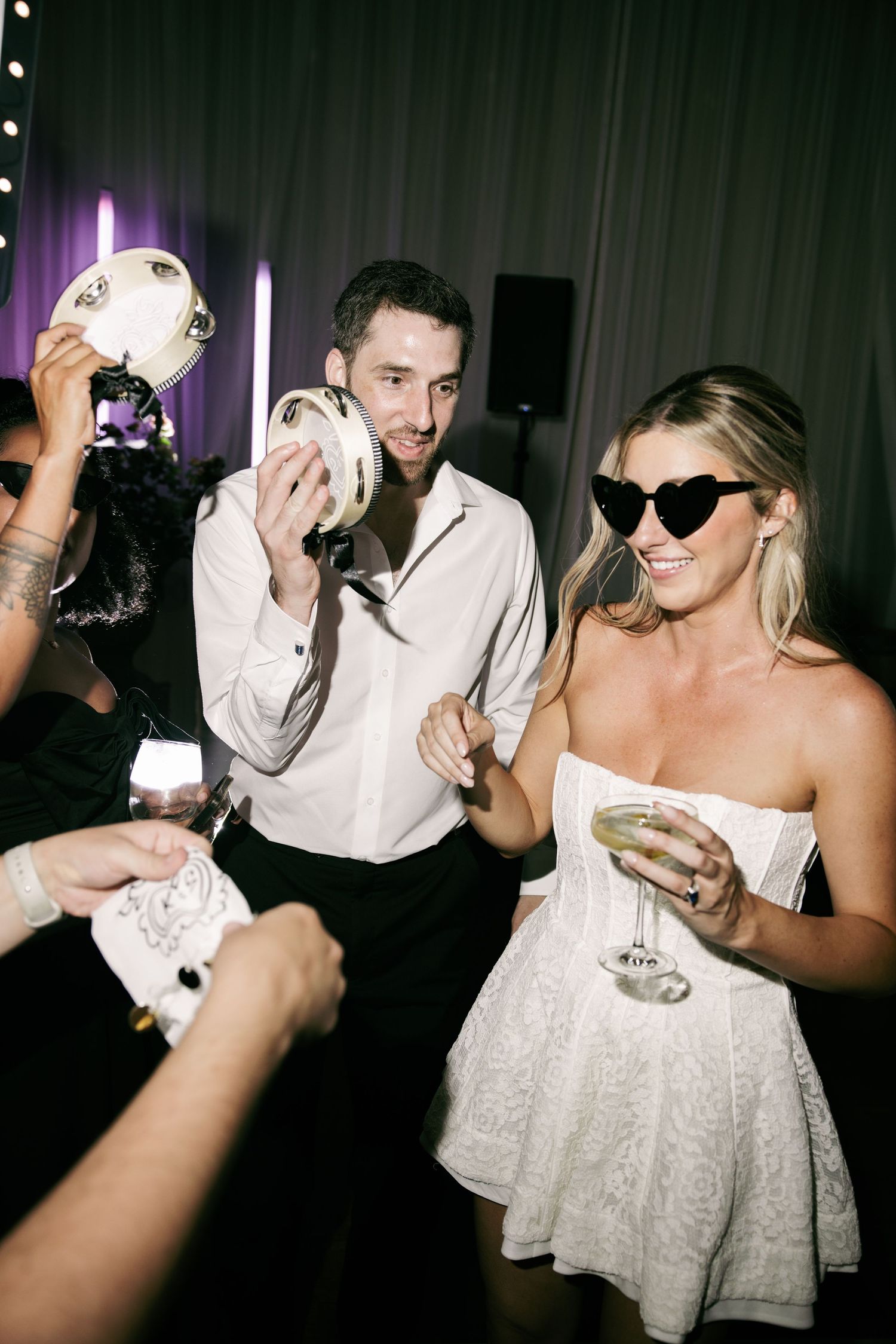 Bride in white strapless mini dress and heart sunglasses celebrating at wedding reception party with guests.