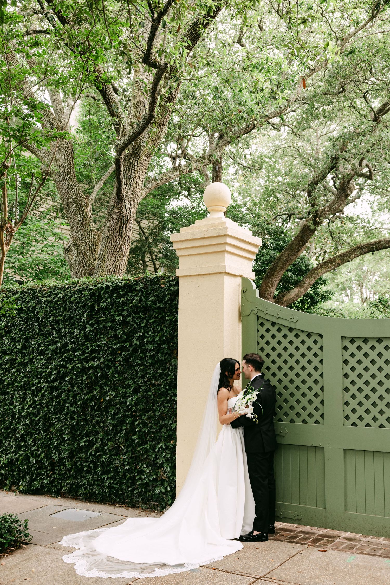 Bride and groom share a romantic kiss by an ivy-covered garden gate surrounded by lush green trees.