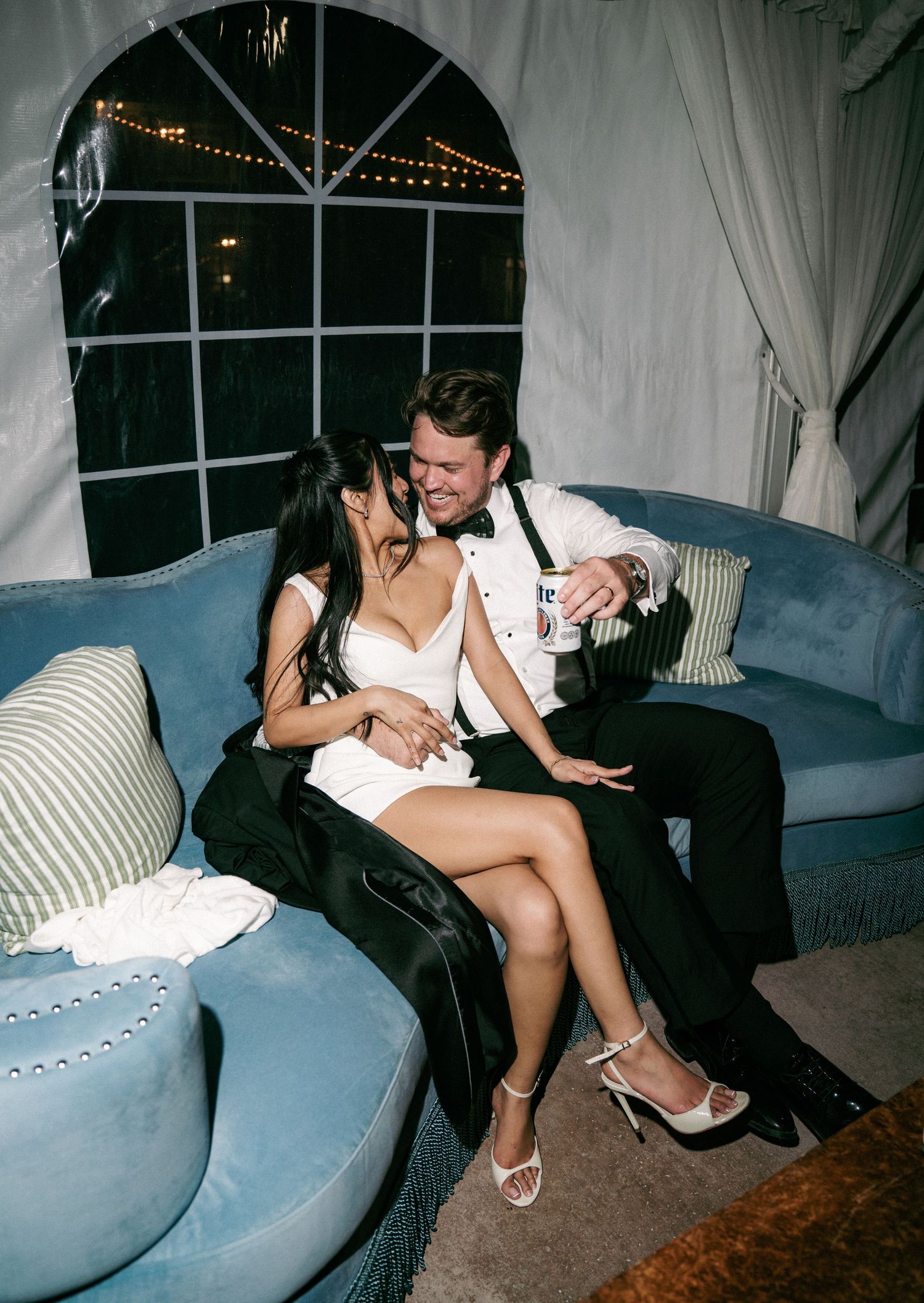 Bride and groom relaxing on blue velvet sofa laughing together at evening wedding reception.