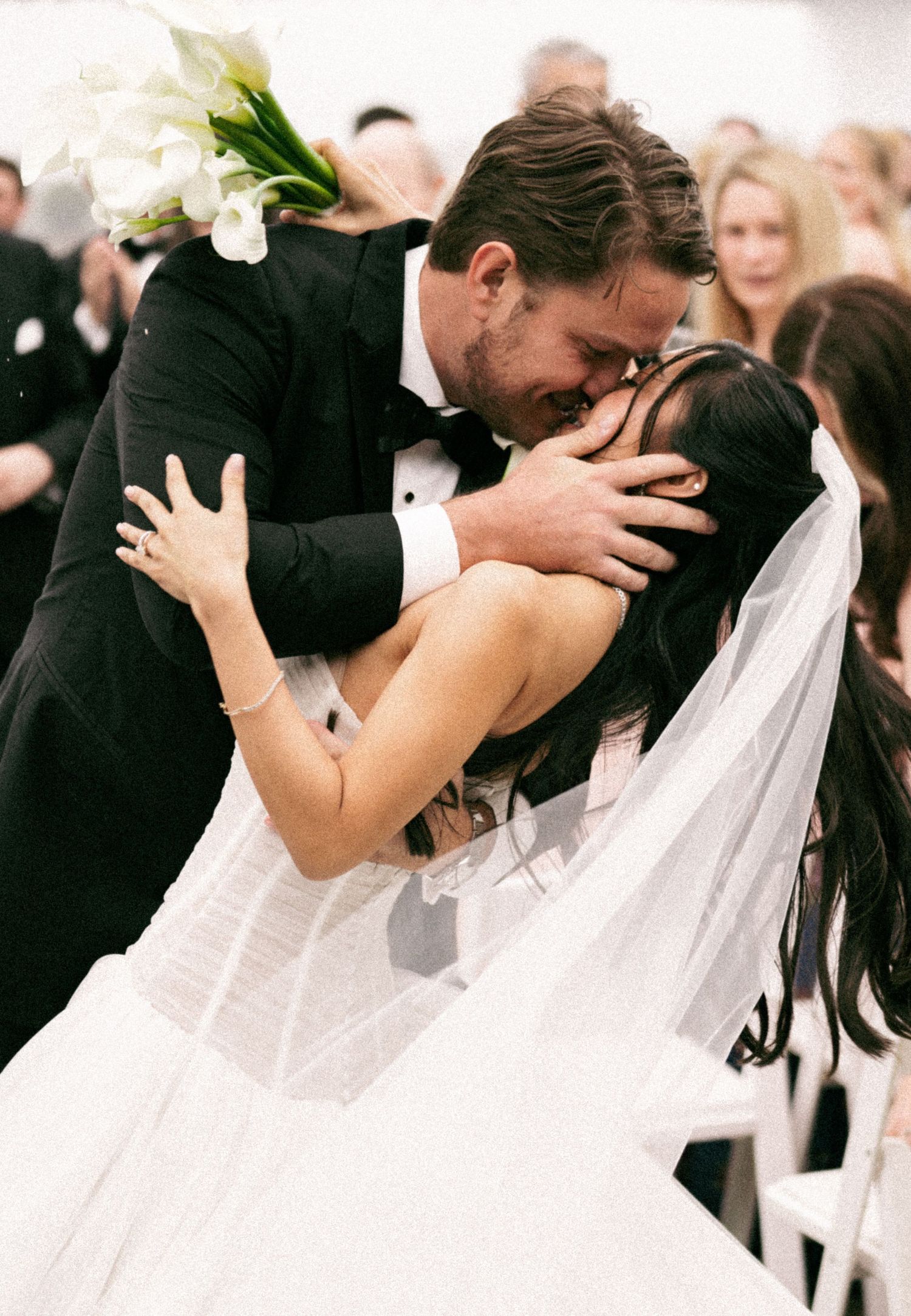 Groom in black tuxedo shares emotional first kiss with bride in white veil during wedding ceremony.