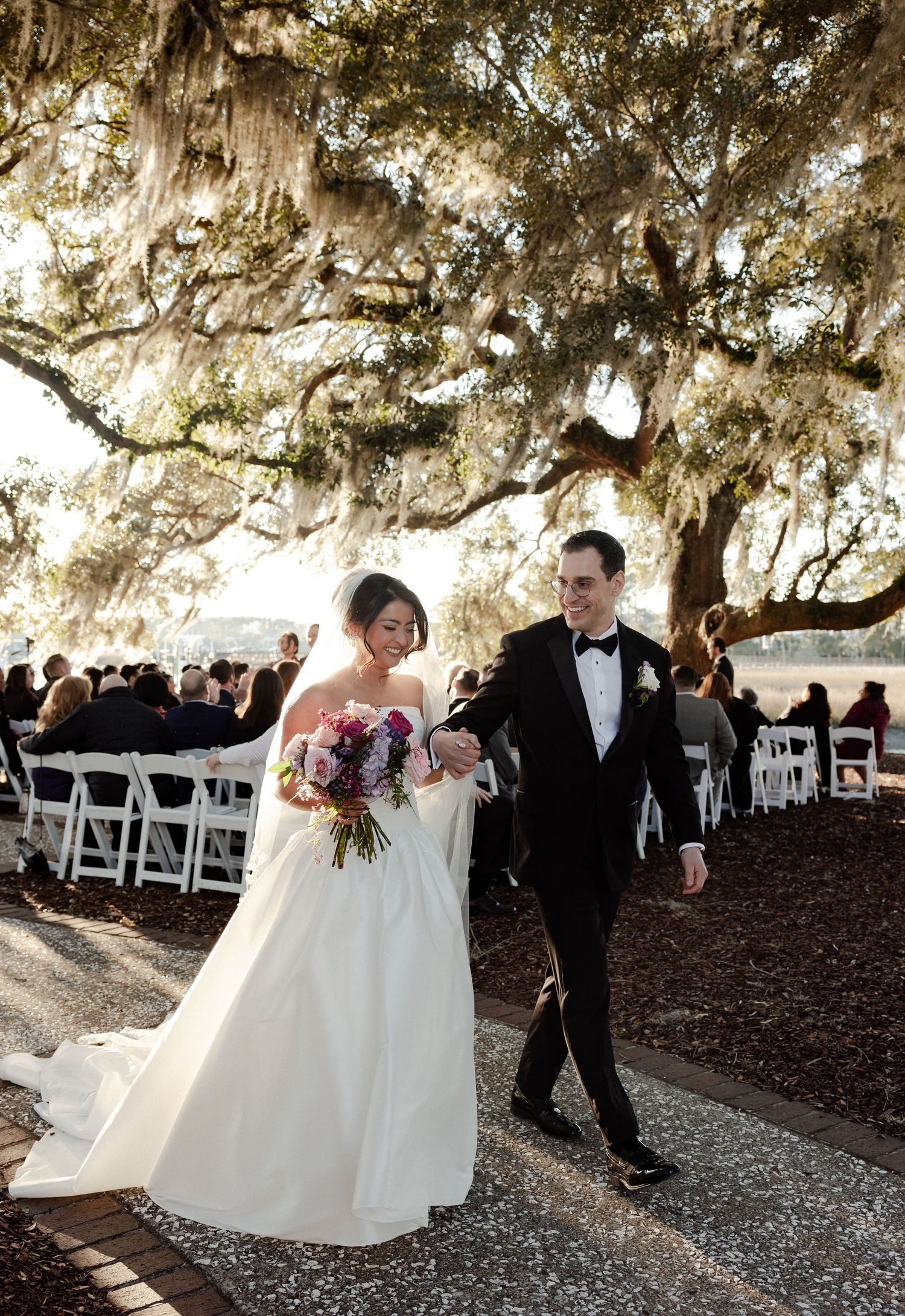 A wedding ceremony under majestic oak trees draped with Spanish moss, with a couple walking down a gravel path.