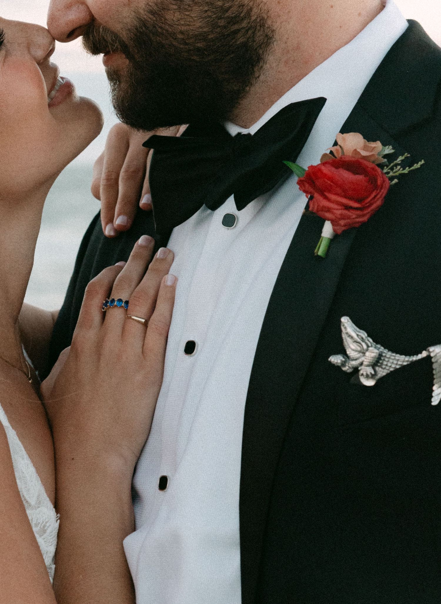 Close-up of groom in black tuxedo with red rose boutonniere and bride wearing sapphire ring.