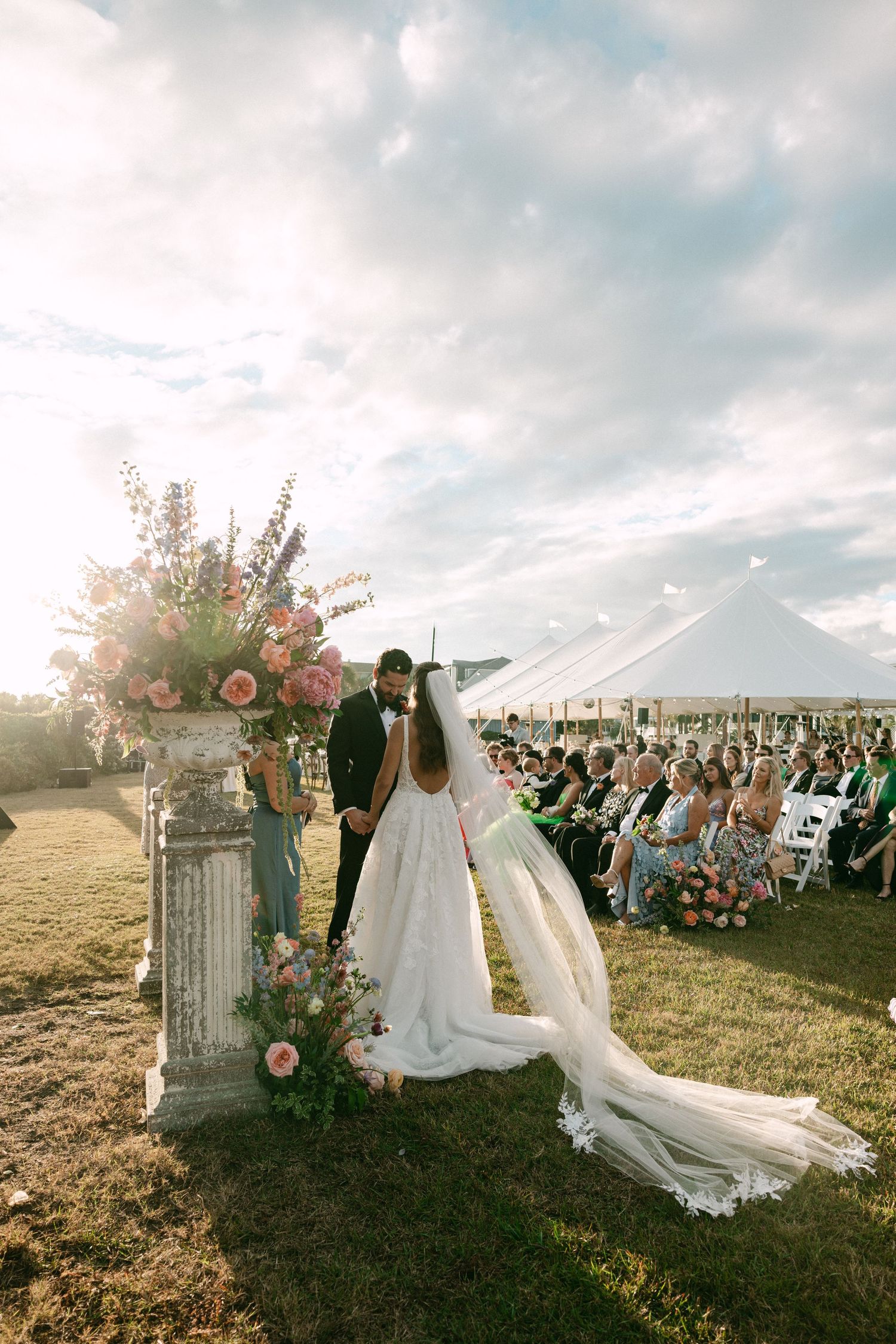 Bride and groom share a kiss at outdoor wedding ceremony beside floral urn with guests seated under white tent.