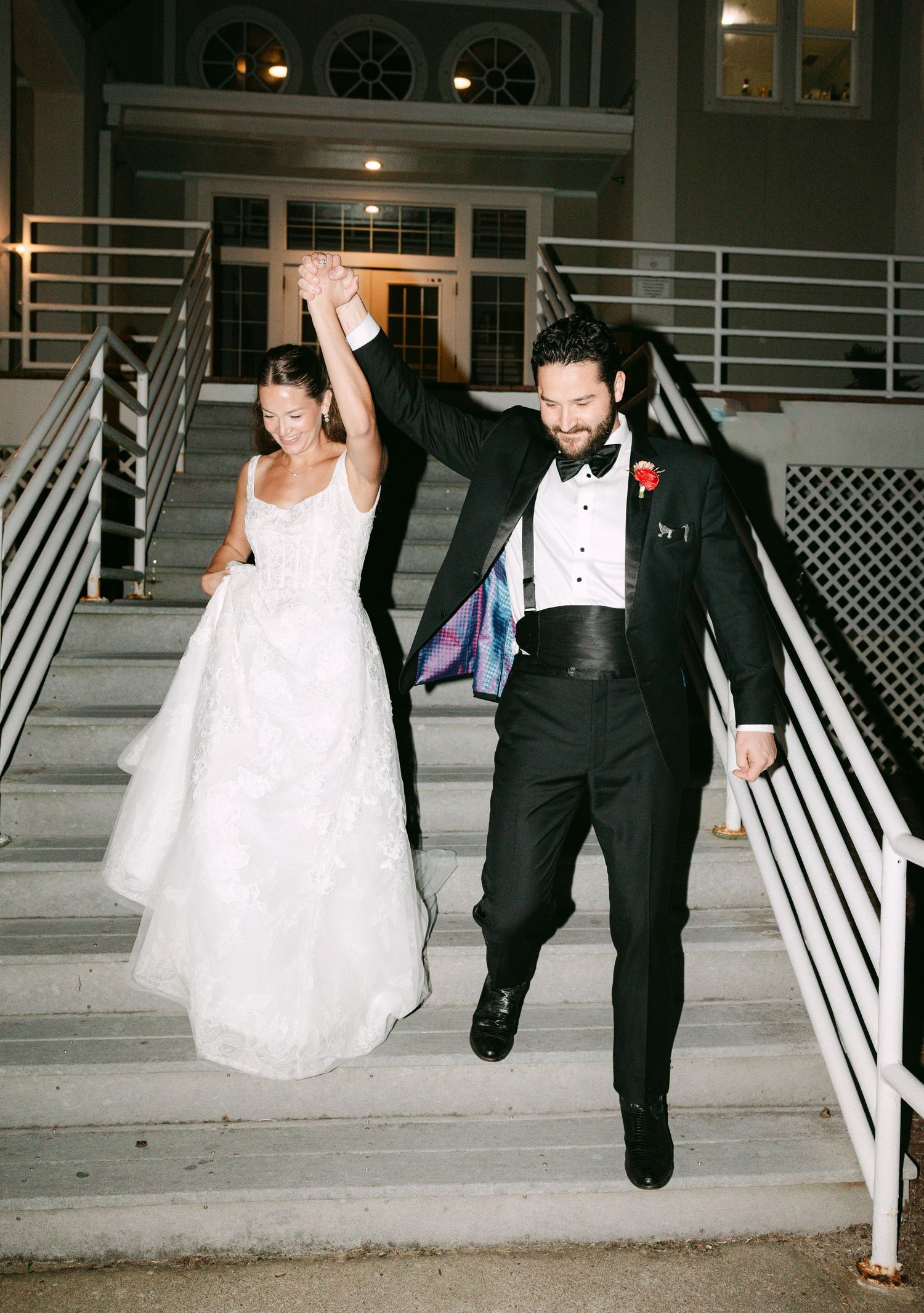 Newlyweds celebrate on staircase at night, groom raising bride's hand in triumph after reception.