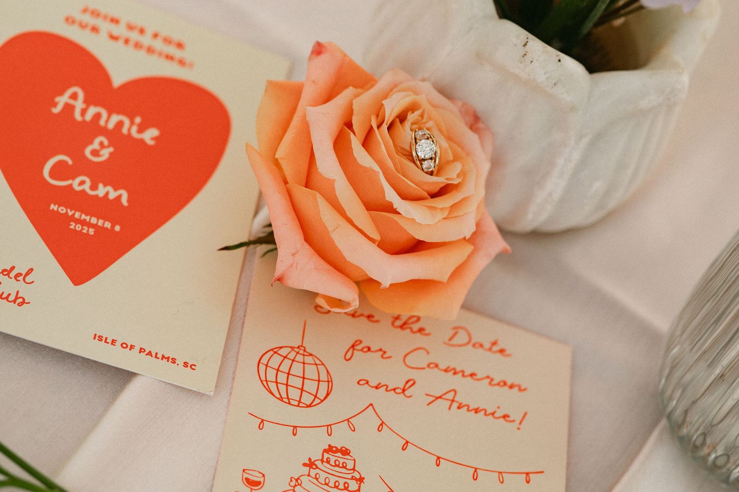 Coral wedding stationery with Annie and Cam save the date cards beside a peach rose with an engagement ring on top.