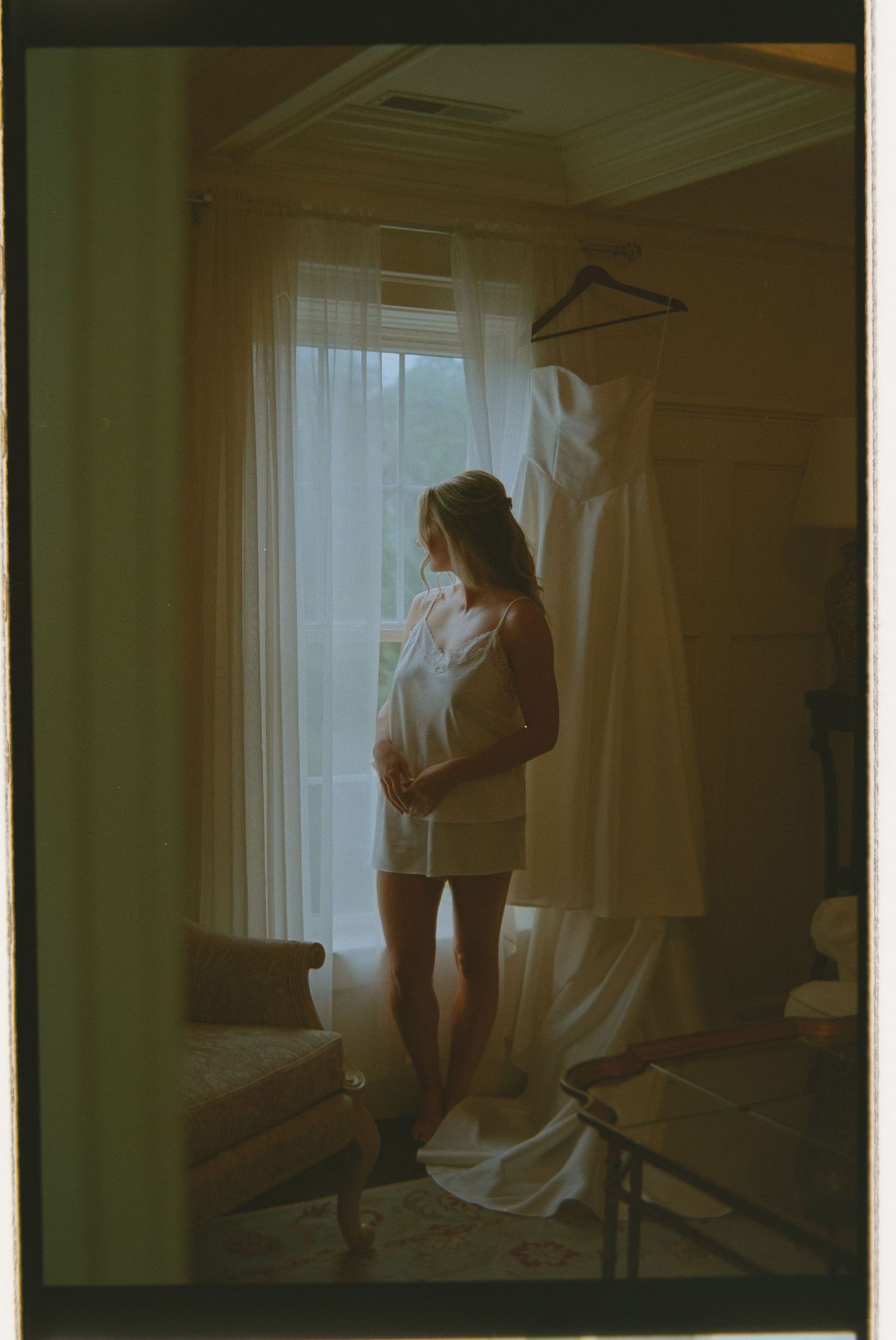 Bride in white slip dress gazes out window beside her hanging wedding gown in softly lit room.