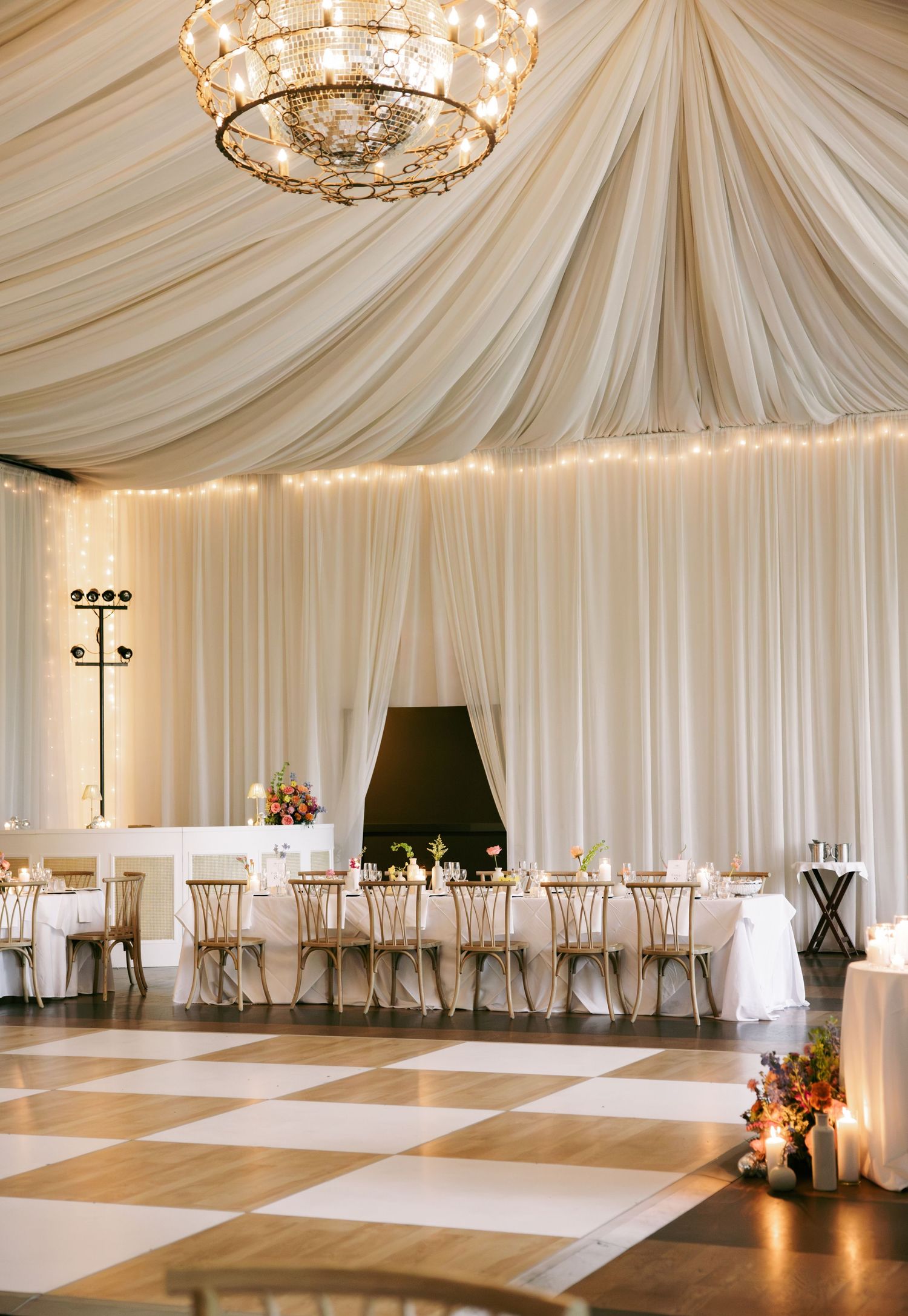Elegant wedding reception with draped white ceiling, chandelier, and checkered dance floor.
