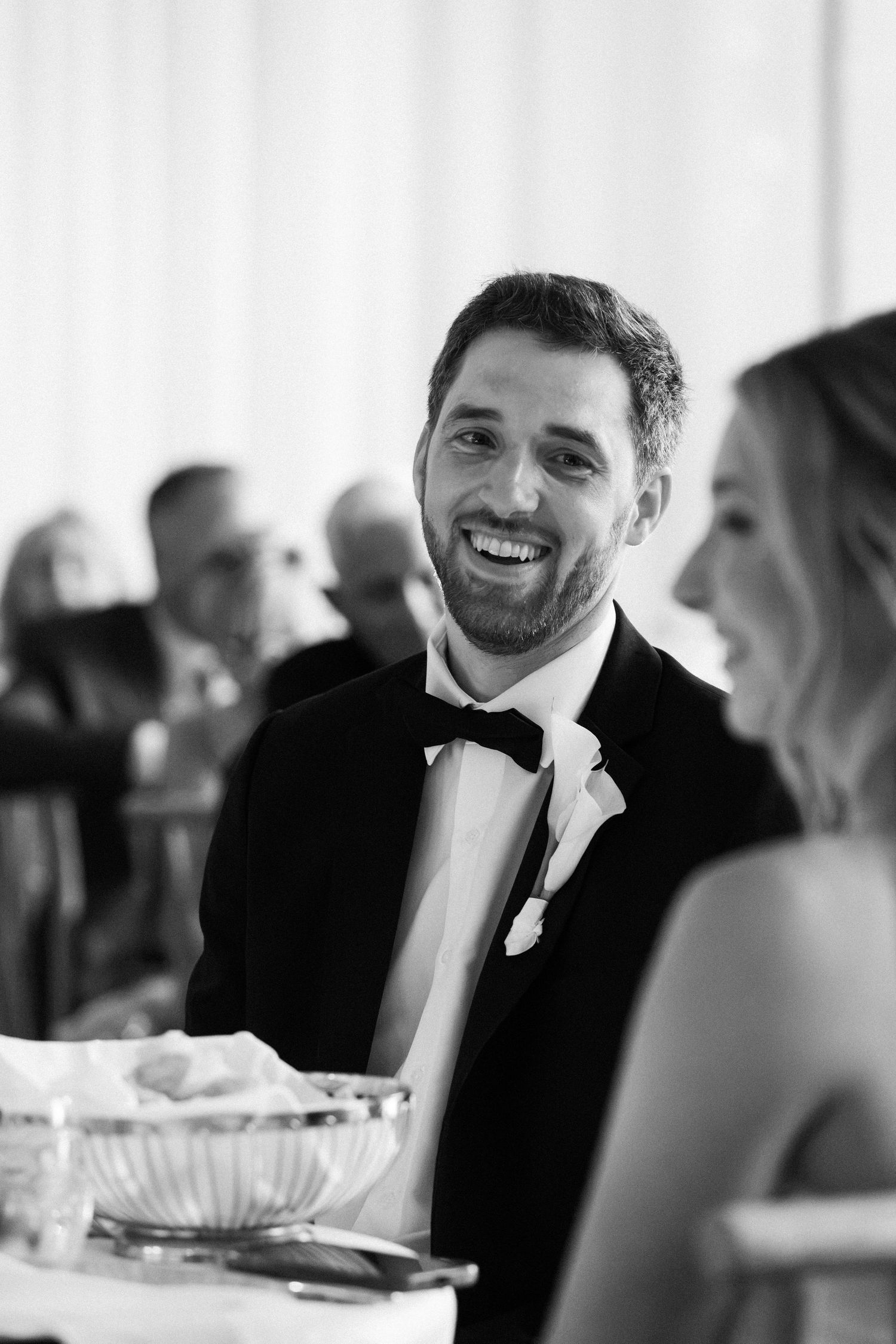 Black and white photo of laughing groom in tuxedo seated at wedding reception dinner table.