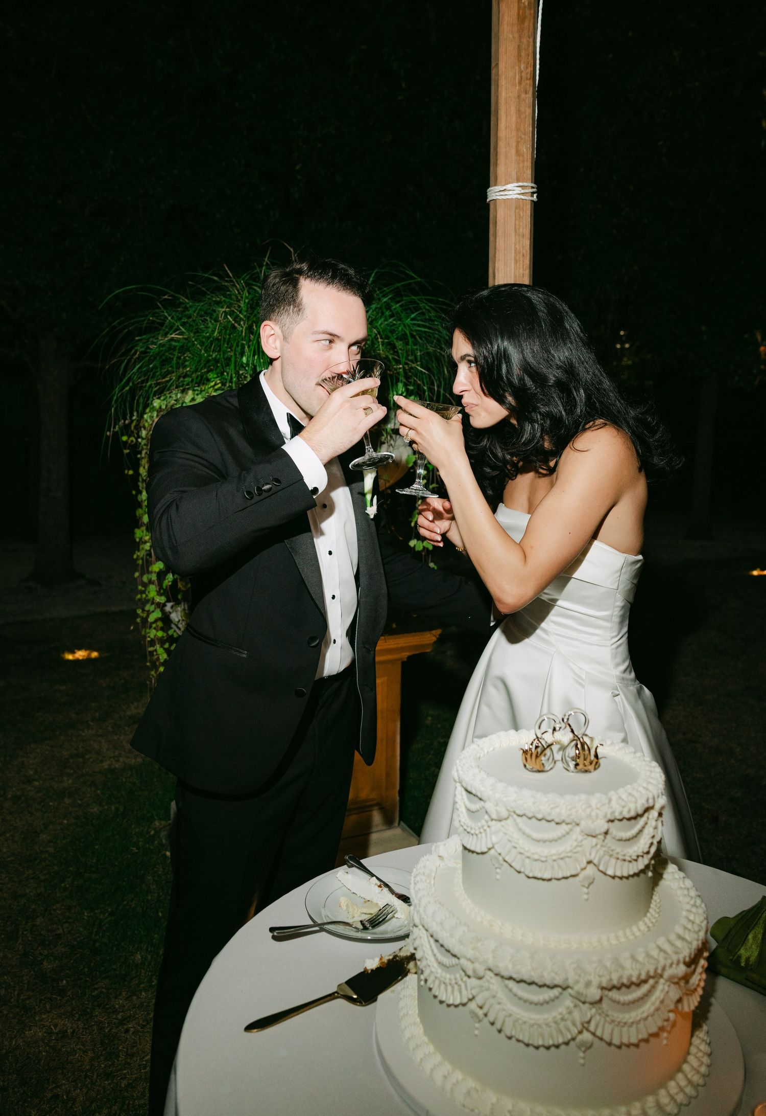 Bride and groom share cake at outdoor wedding reception at night, dressed in white gown and black tuxedo.