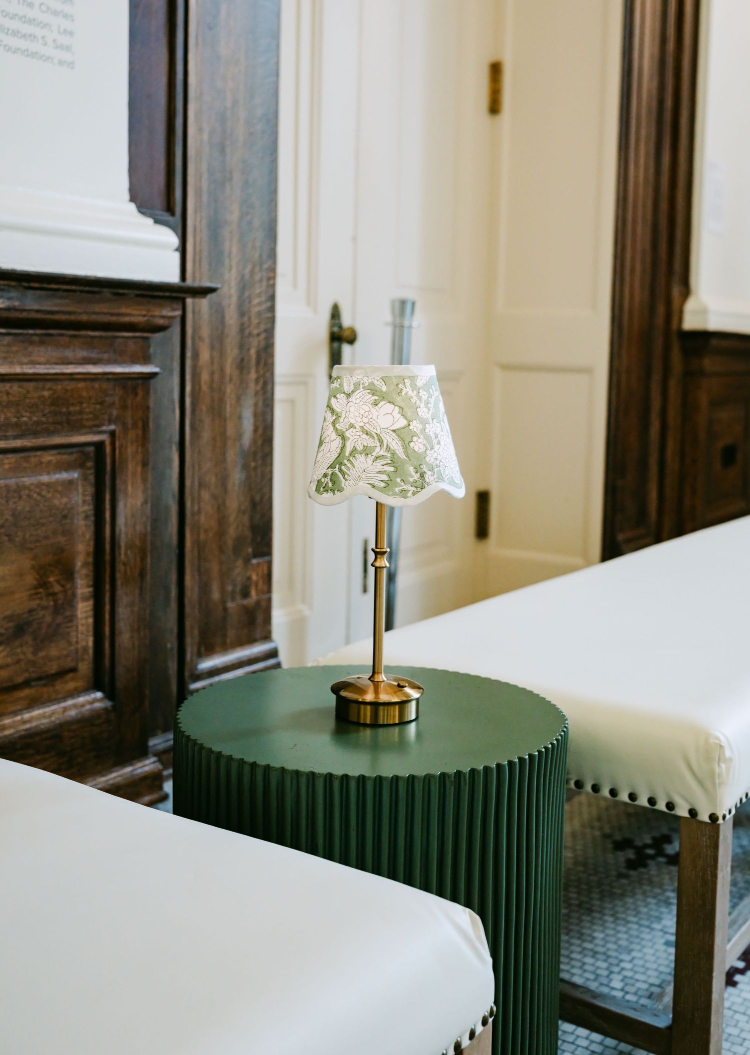 Green fluted side table with brass lamp and floral shade between white upholstered benches in elegant interior.