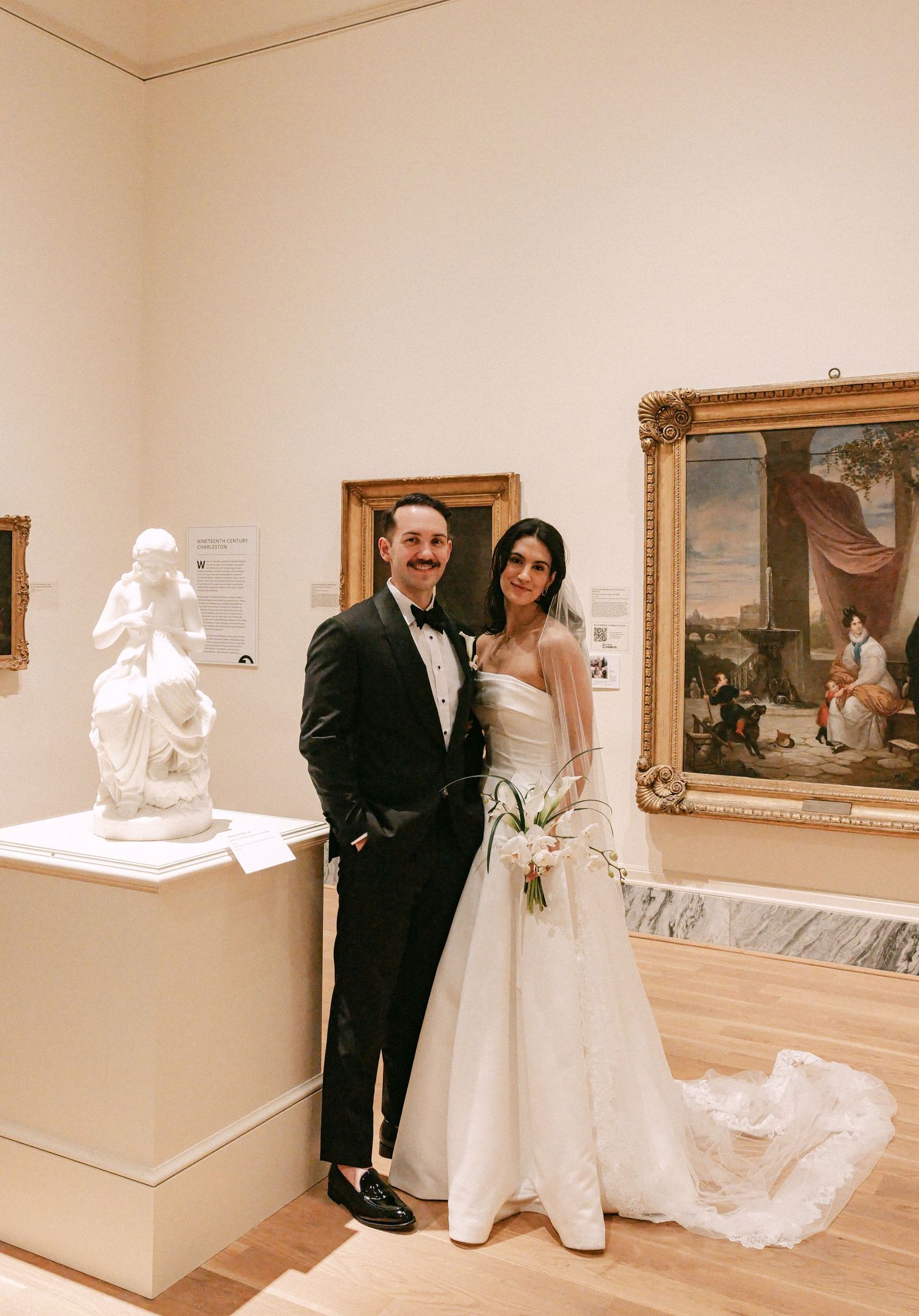 Bride and groom pose in wedding attire beside sculpture and painting in art museum.