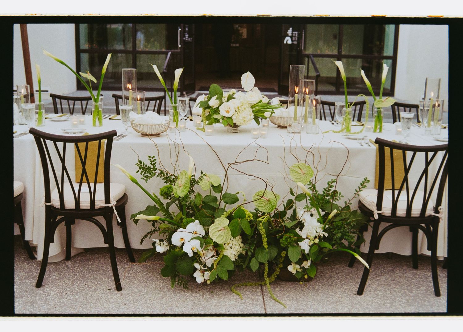 Wedding sweetheart table with white floral centerpiece featuring greenery, roses, and calla lilies on white linen.