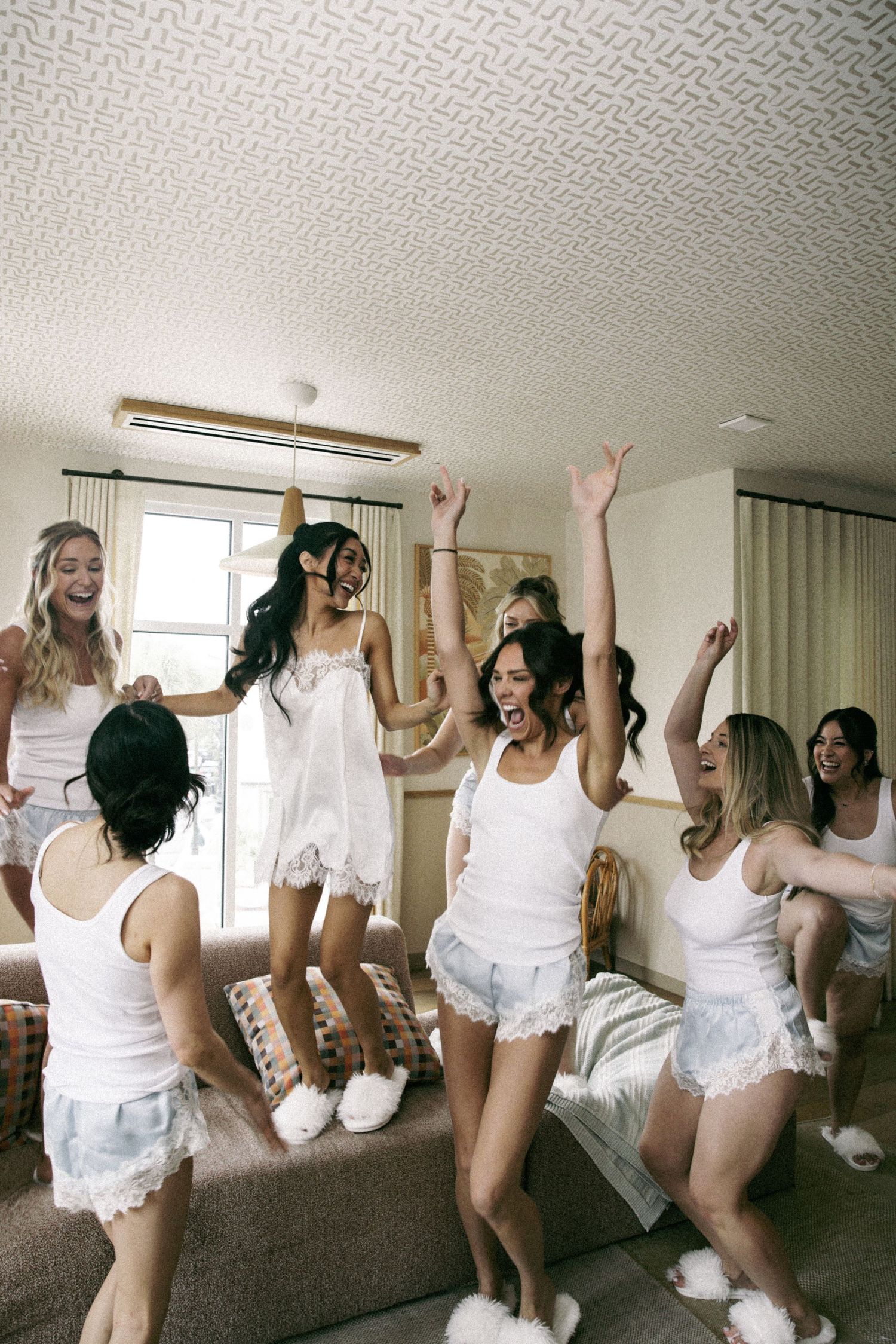 A group of women in white pajamas dancing and celebrating together in a hotel room during a bachelorette party.