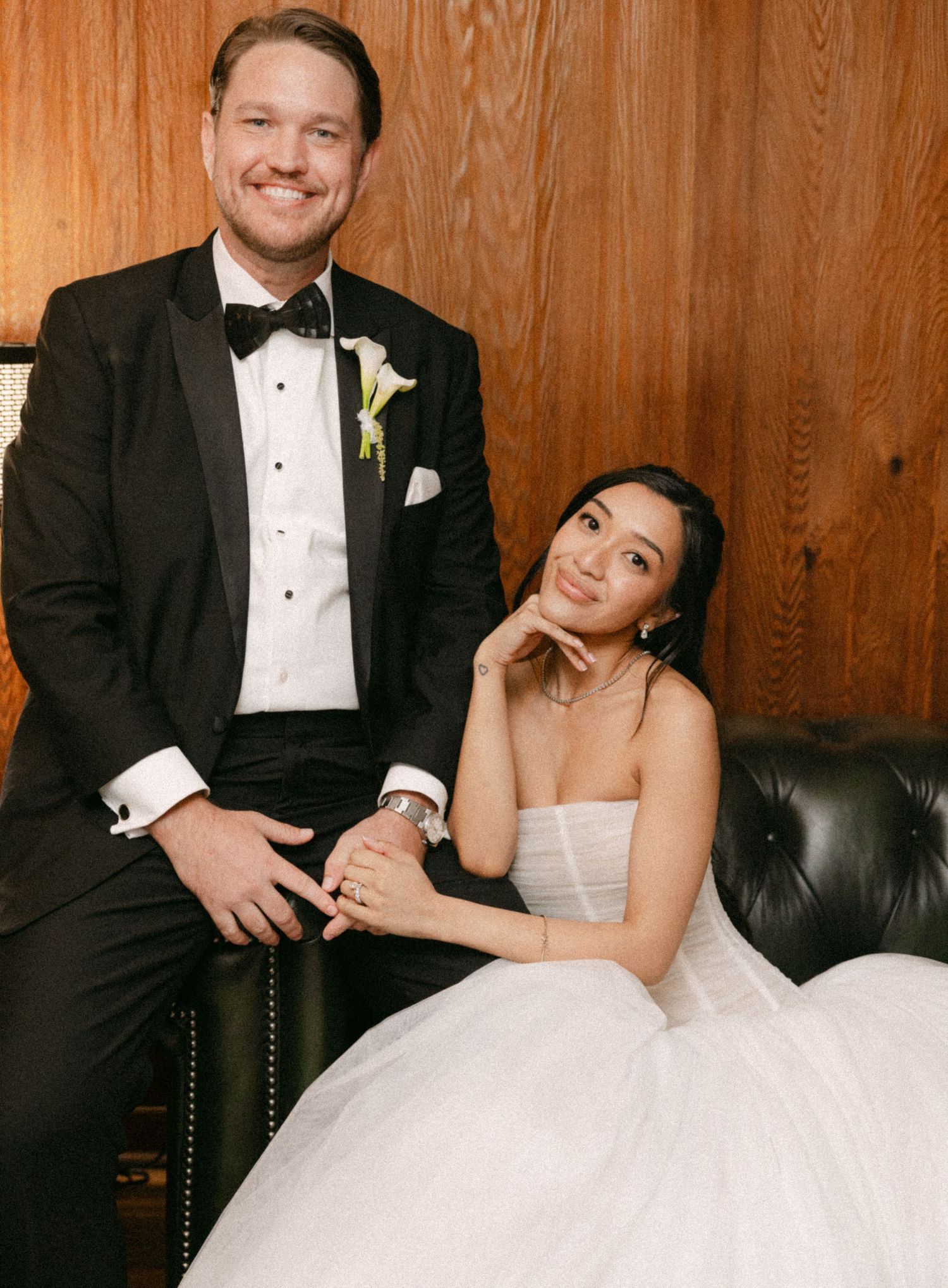 Bride in strapless white ball gown and groom in black tuxedo pose together on leather sofa at wedding.